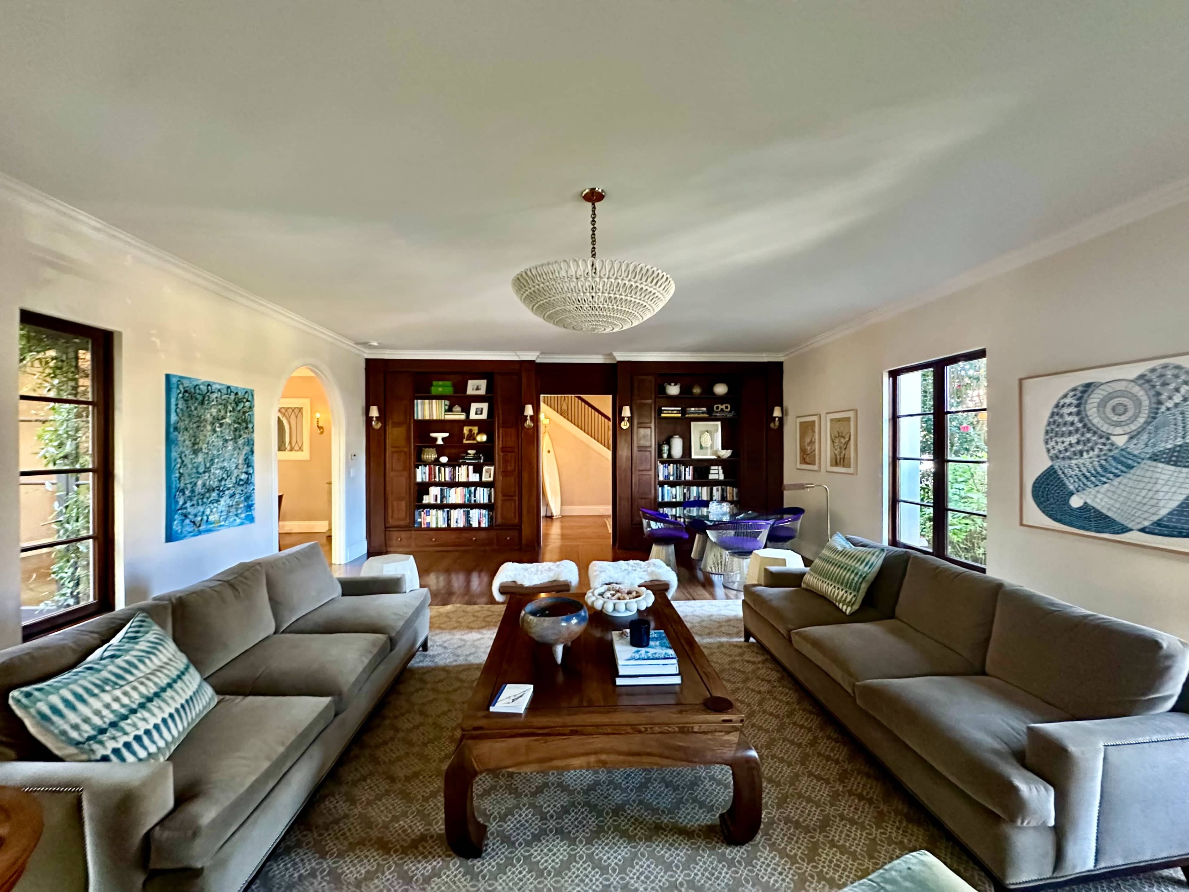 The living room features two sofas facing a wooden coffee table, with bookshelves and artwork lining the walls, and a staircase visible in the background.