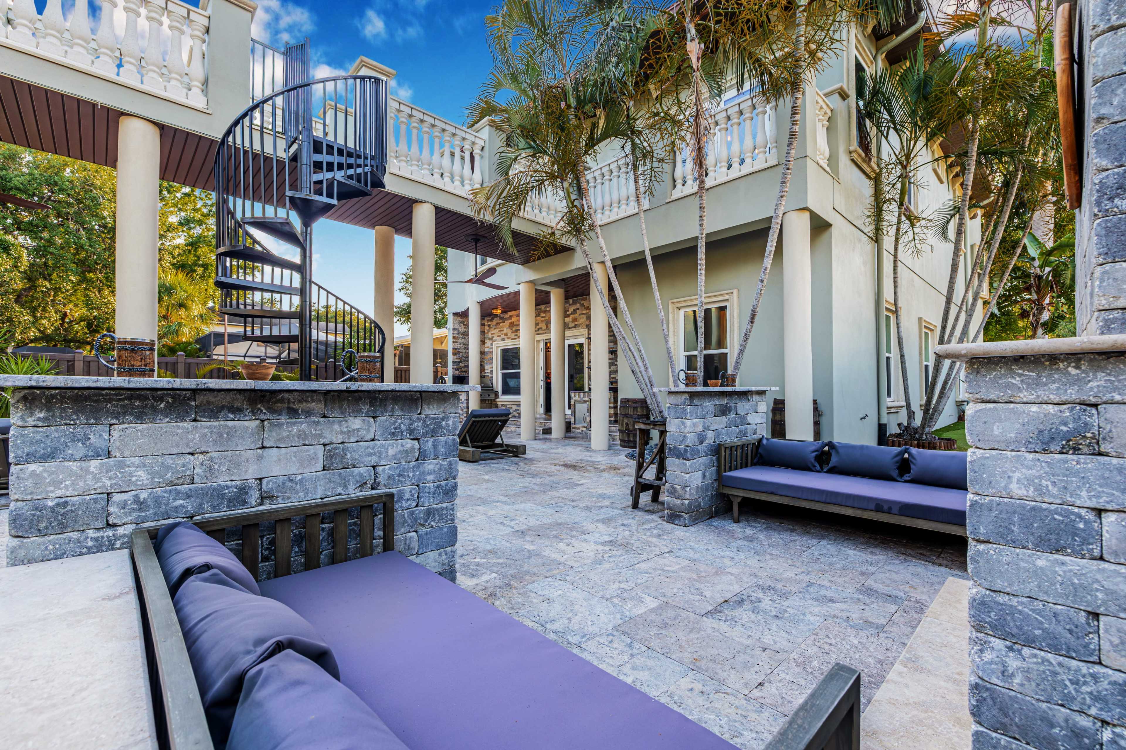The image shows a modern outdoor patio area with spiral stairs leading to a second level and seating arrangements made of dark wood and stone.