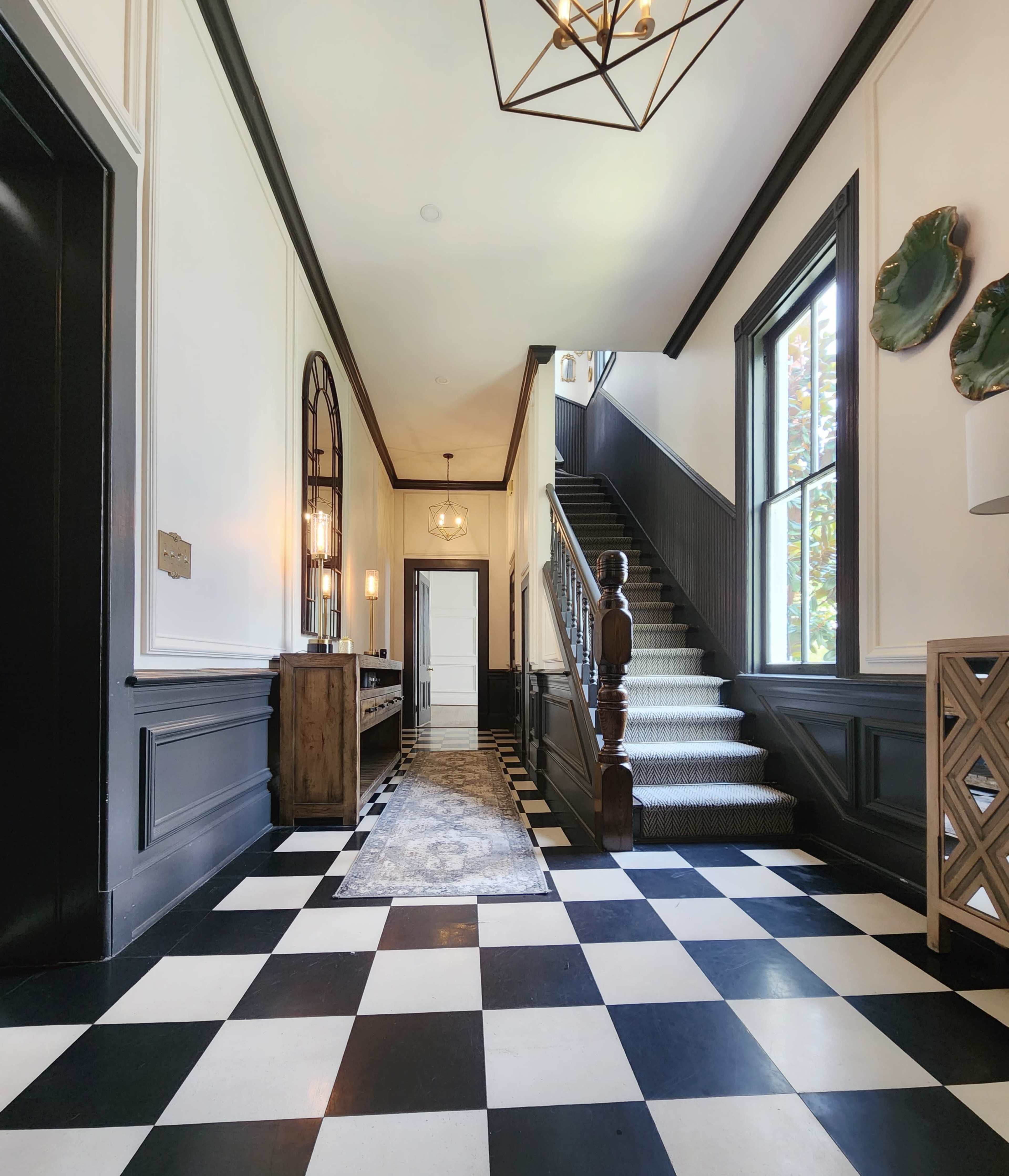 A hallway with a checkered black and white floor, a staircase on the right, and decorative elements including a chandelier and wall decor.