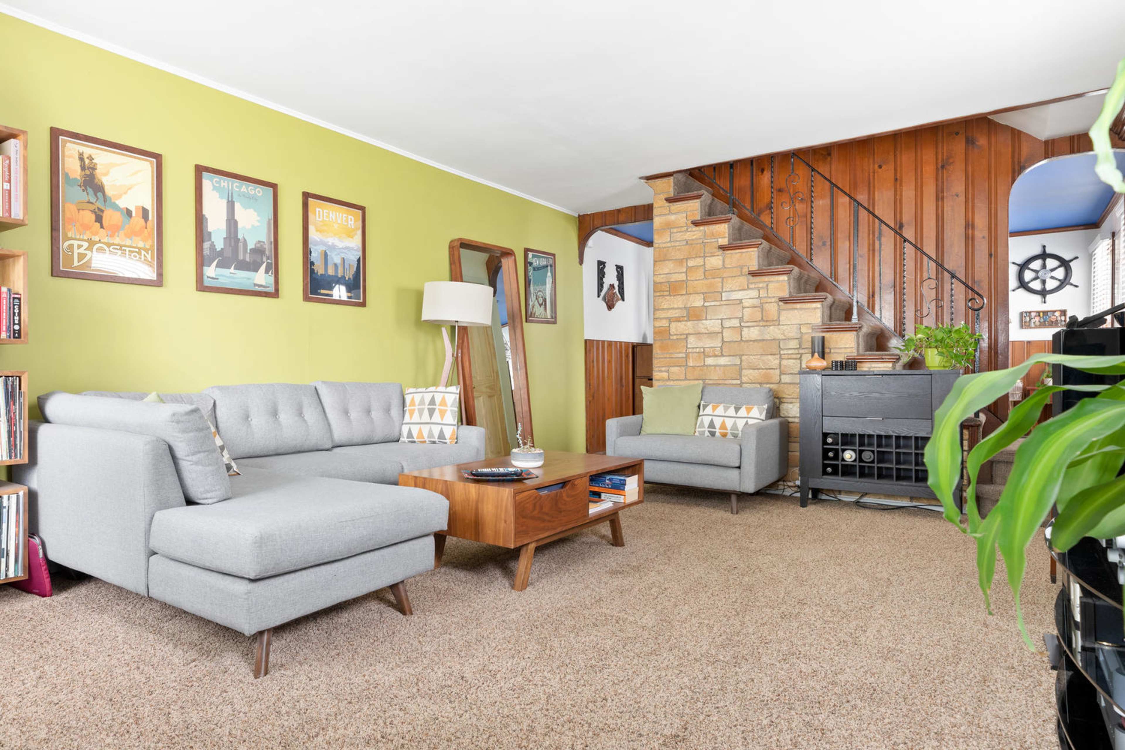 A living room with a gray sectional sofa, a wooden coffee table, and a staircase leading to an upper level, all surrounded by green and wooden walls.