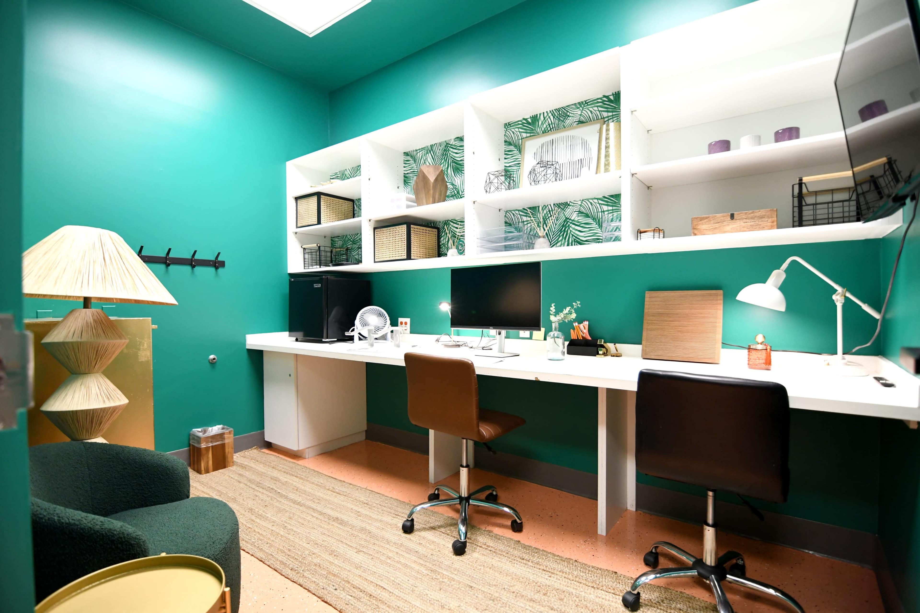 The image shows a modern office space with vibrant green walls, white shelves holding decorative items, two desks with computers, and a comfortable chair in the corner.