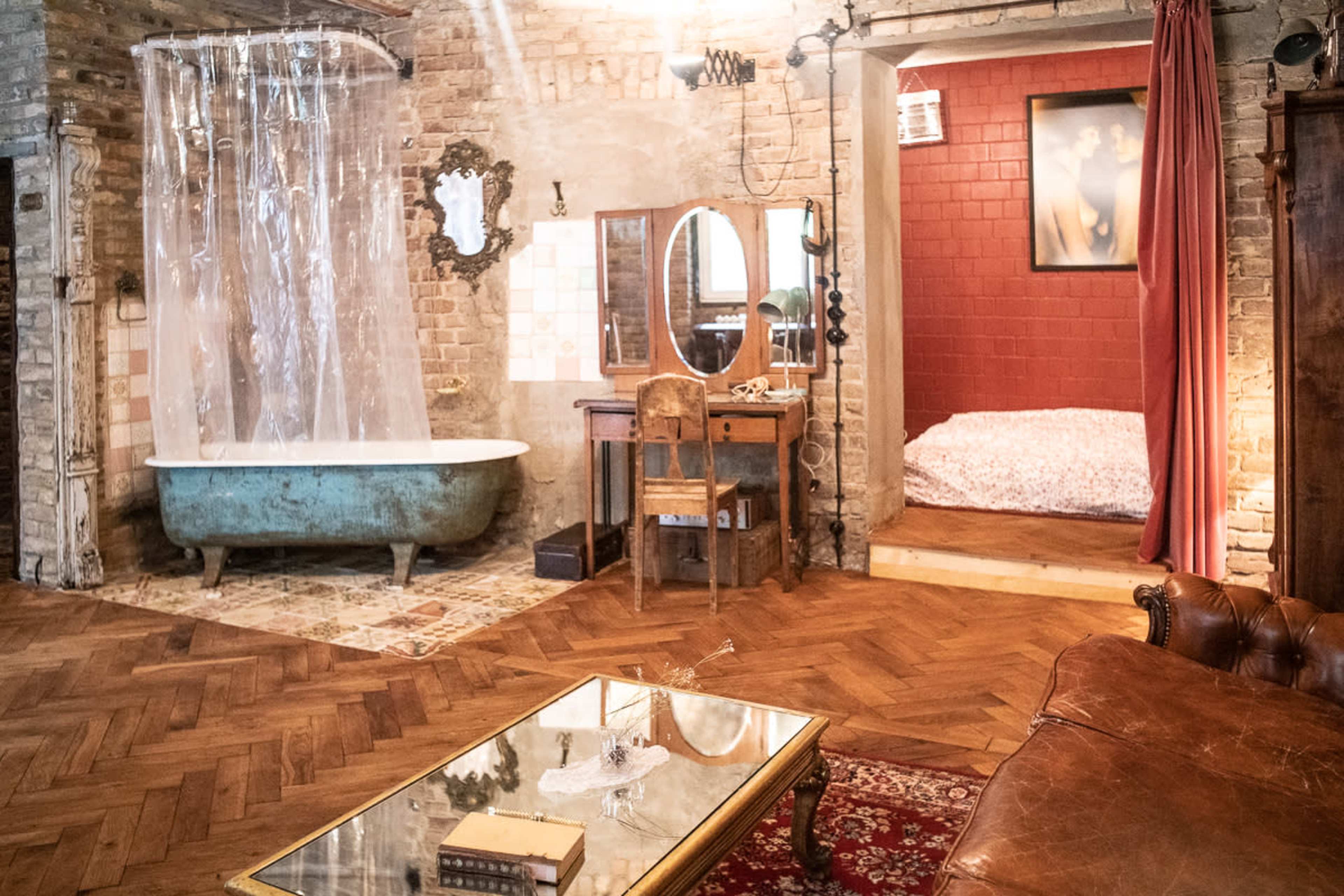A rustic room with a vintage bathtub, a wooden vanity, a brown leather couch, a coffee table, and a bed in a separate alcove.