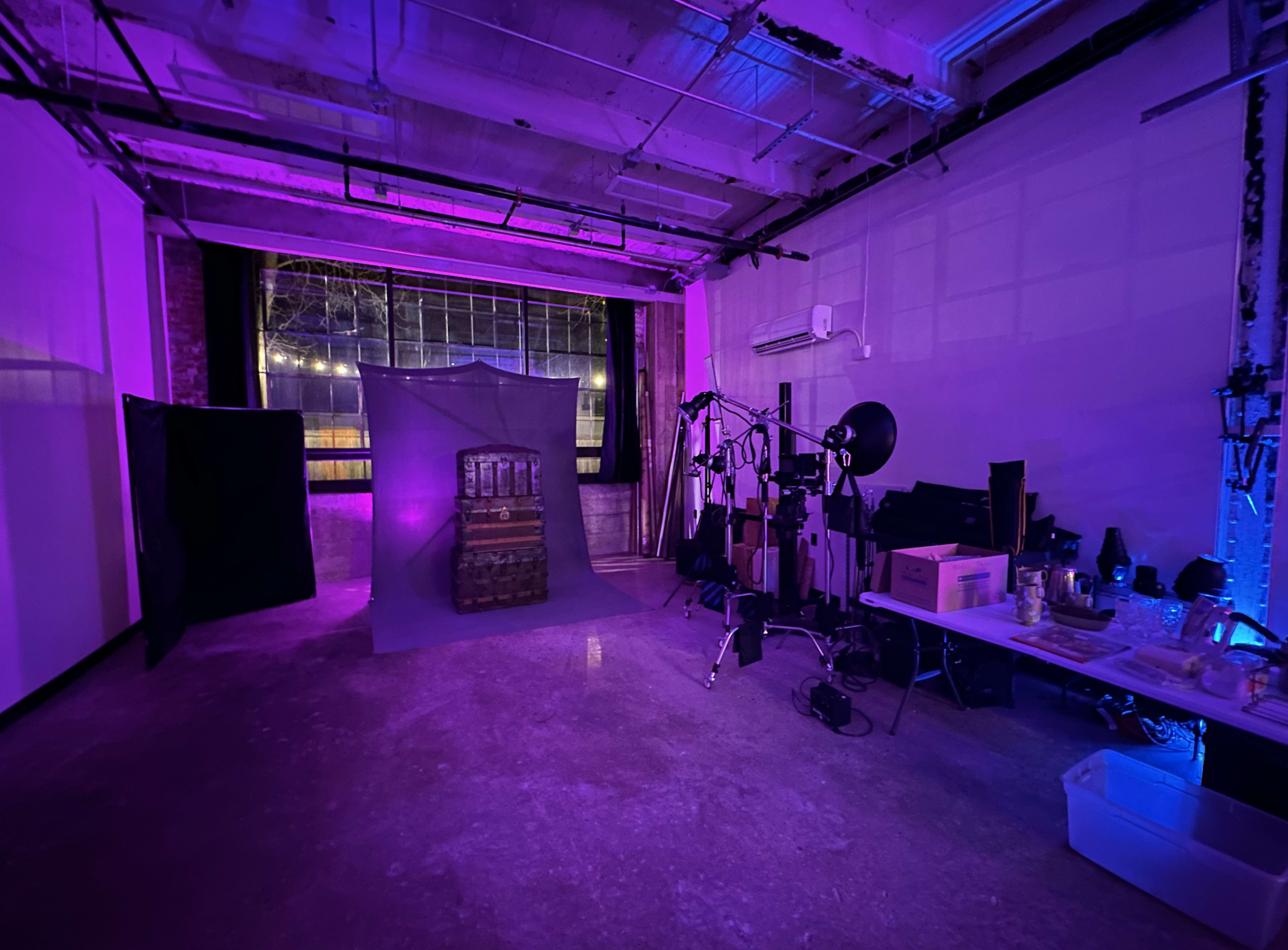 An industrial studio space with purple lighting, a backdrop with a stacked barrel, and various photography equipment arranged nearby.