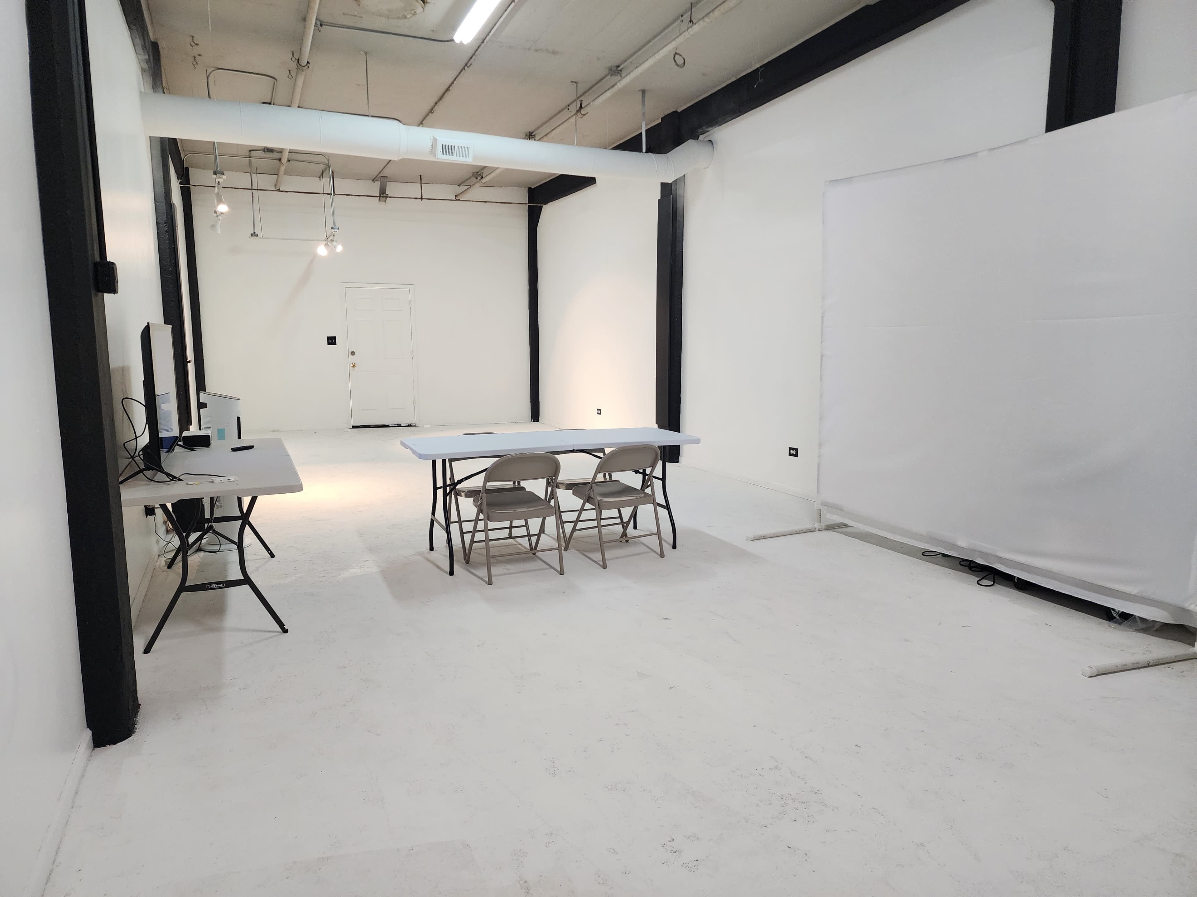 The image shows a minimalist, empty studio space featuring a table with chairs, a computer workstation, and a large white backdrop.