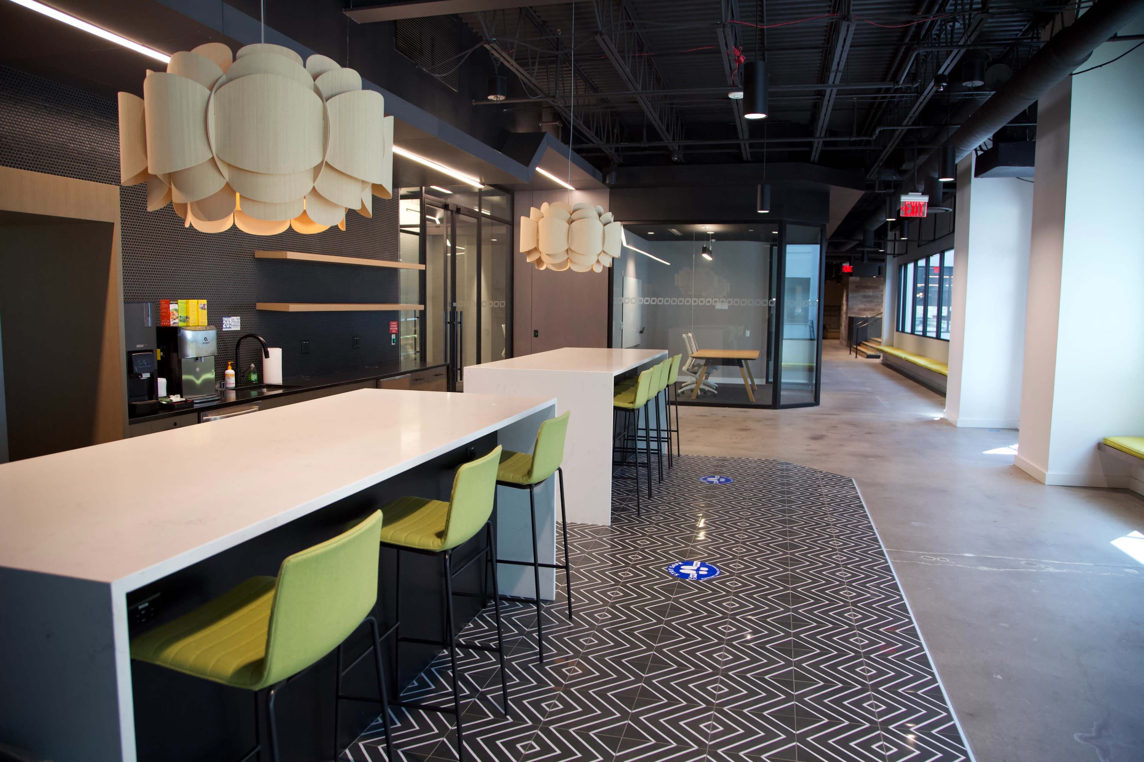 The image shows a modern office break room with a long white countertop, green bar stools, and geometric patterned flooring.