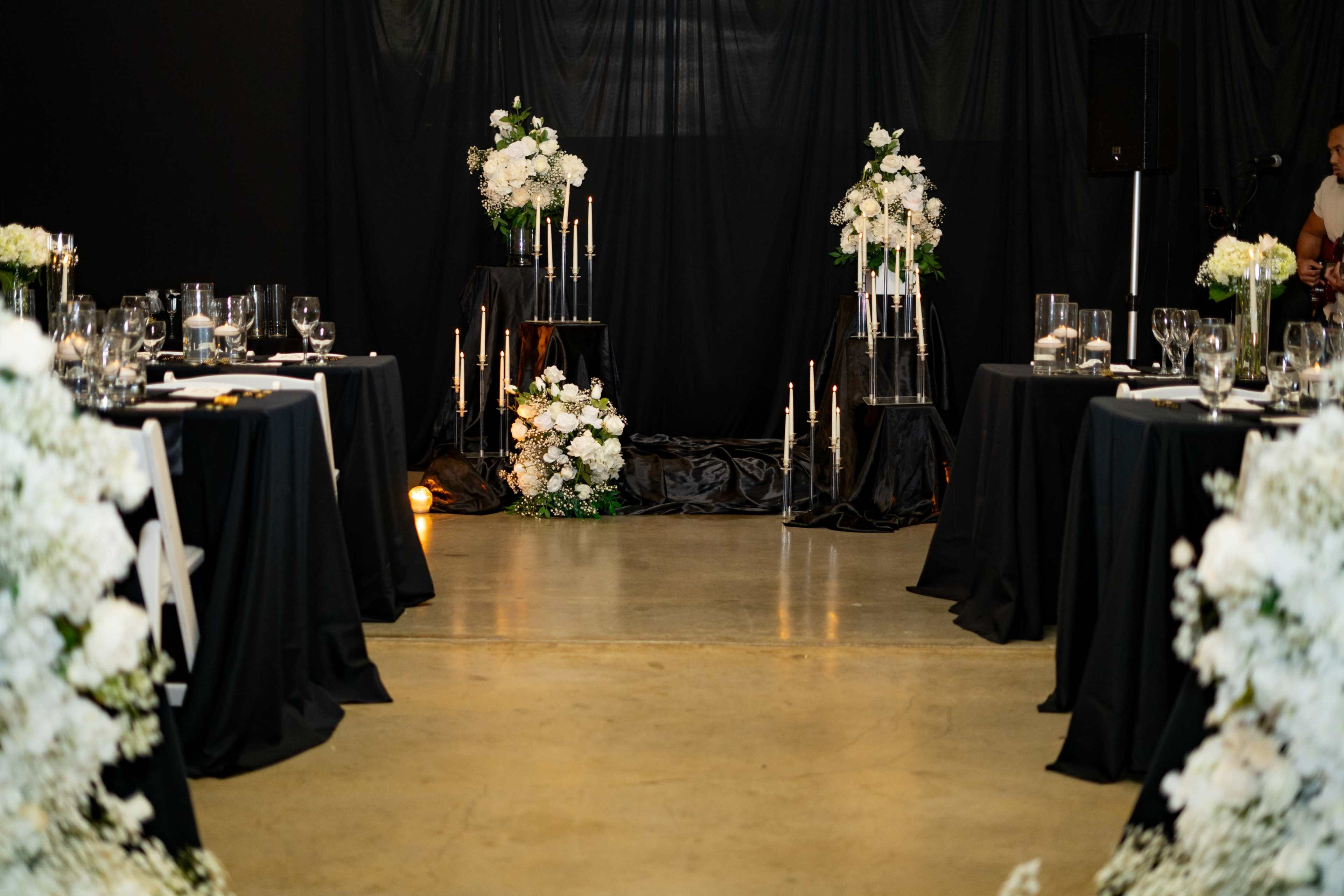 The image shows a decorated event space with black tables adorned with white floral arrangements and candles, creating an elegant atmosphere.