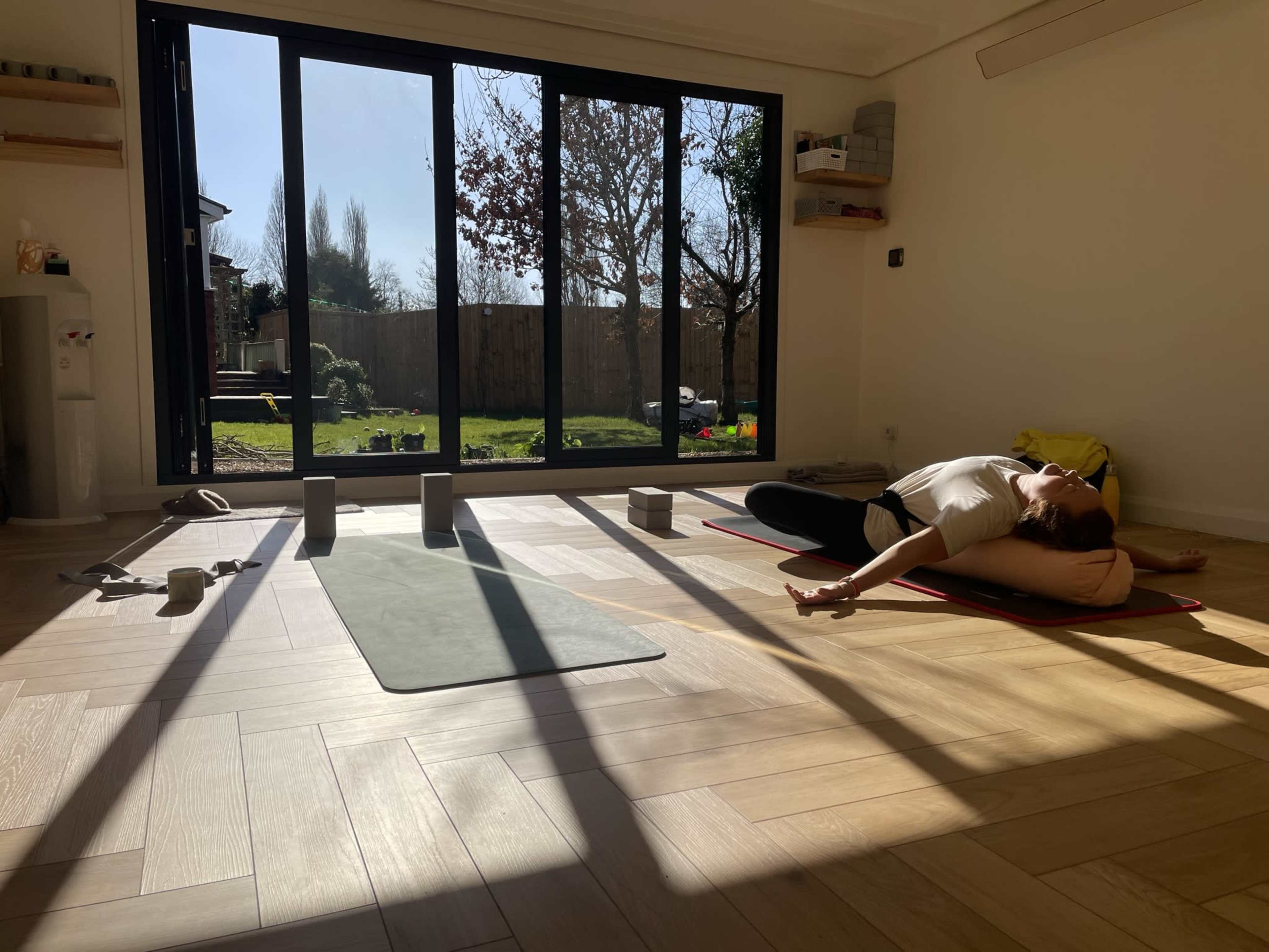 A person practices yoga on a mat in a sunlit room with large glass doors opening to a garden.