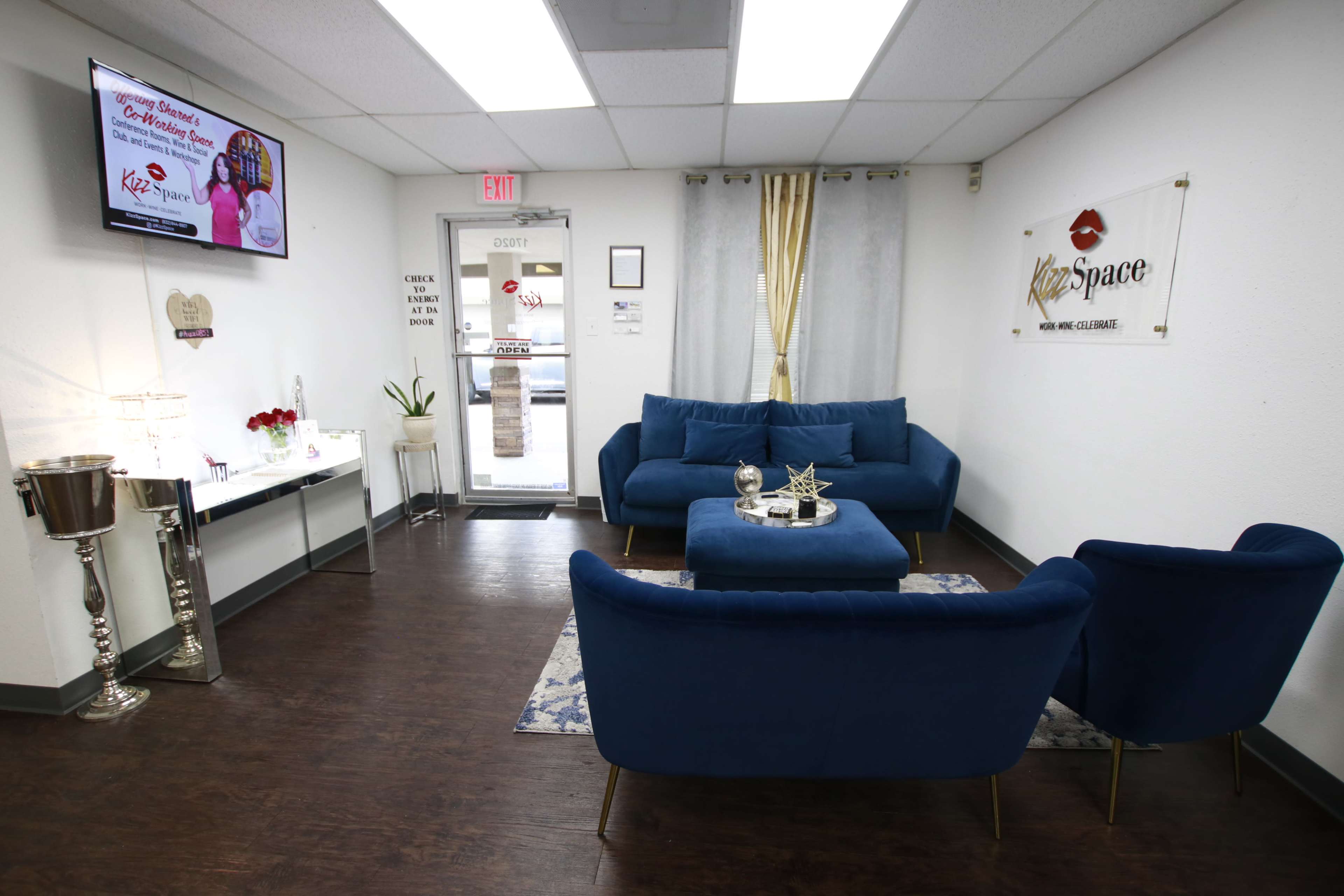 The image shows a modern waiting area with blue upholstered furniture, a decorative table, and a glass door leading outside.