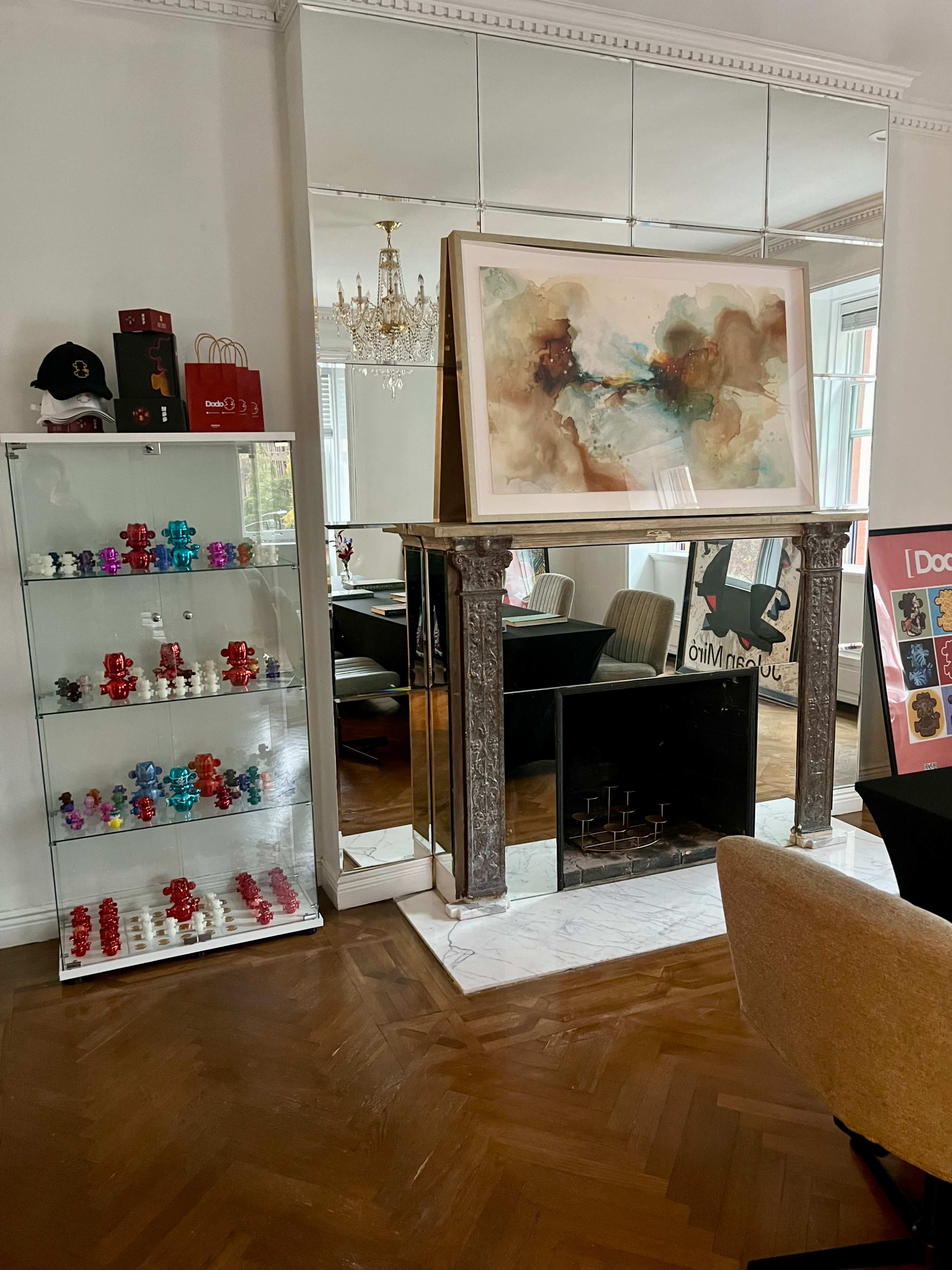 The image shows a room with a mirrored wall featuring a fireplace, a large abstract painting above it, a display case with colorful figurines, and a dining area in the background.