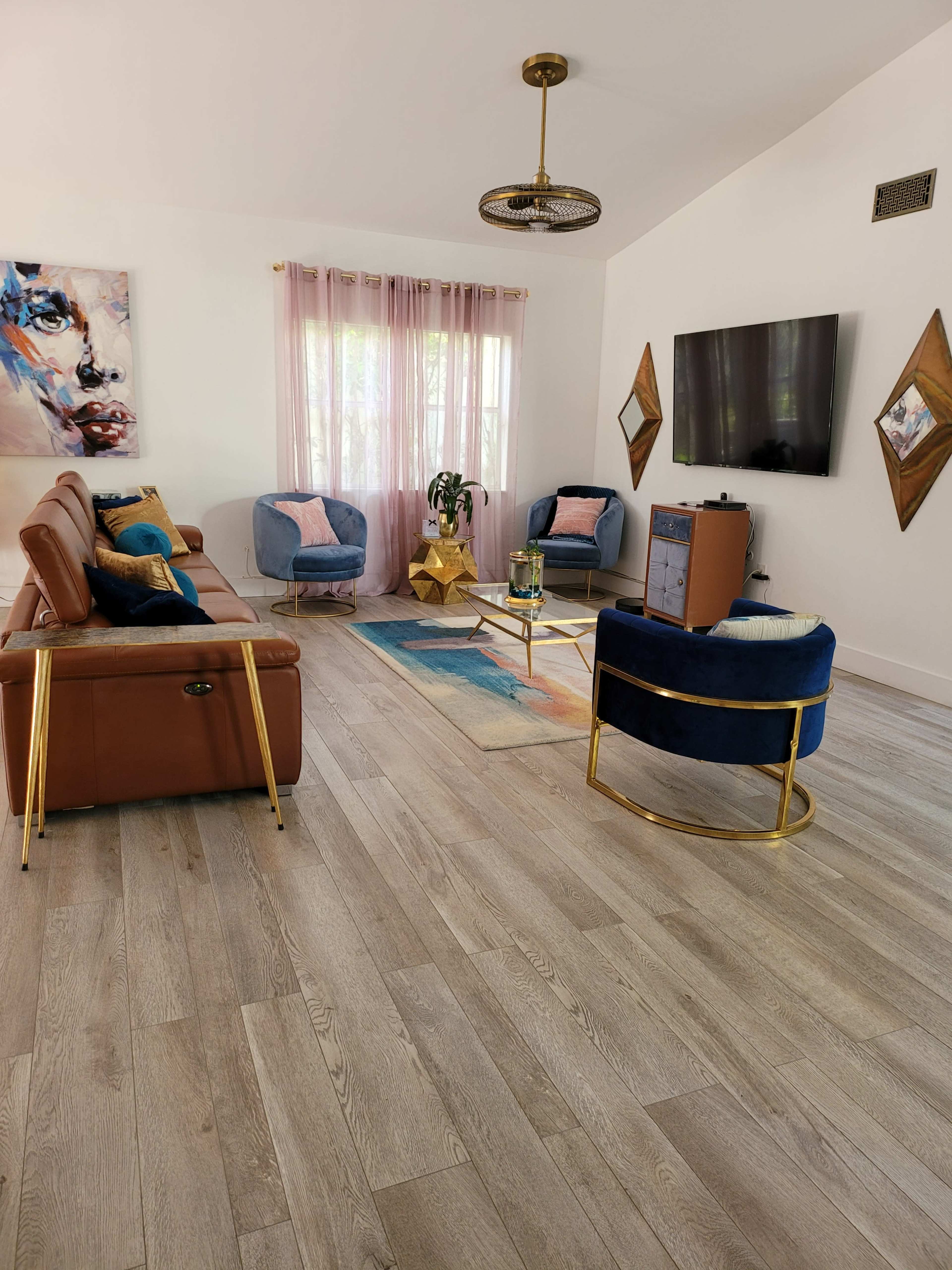 The living room features a mix of brown leather and blue upholstered seating, a gold coffee table, and large windows with sheer pink curtains.