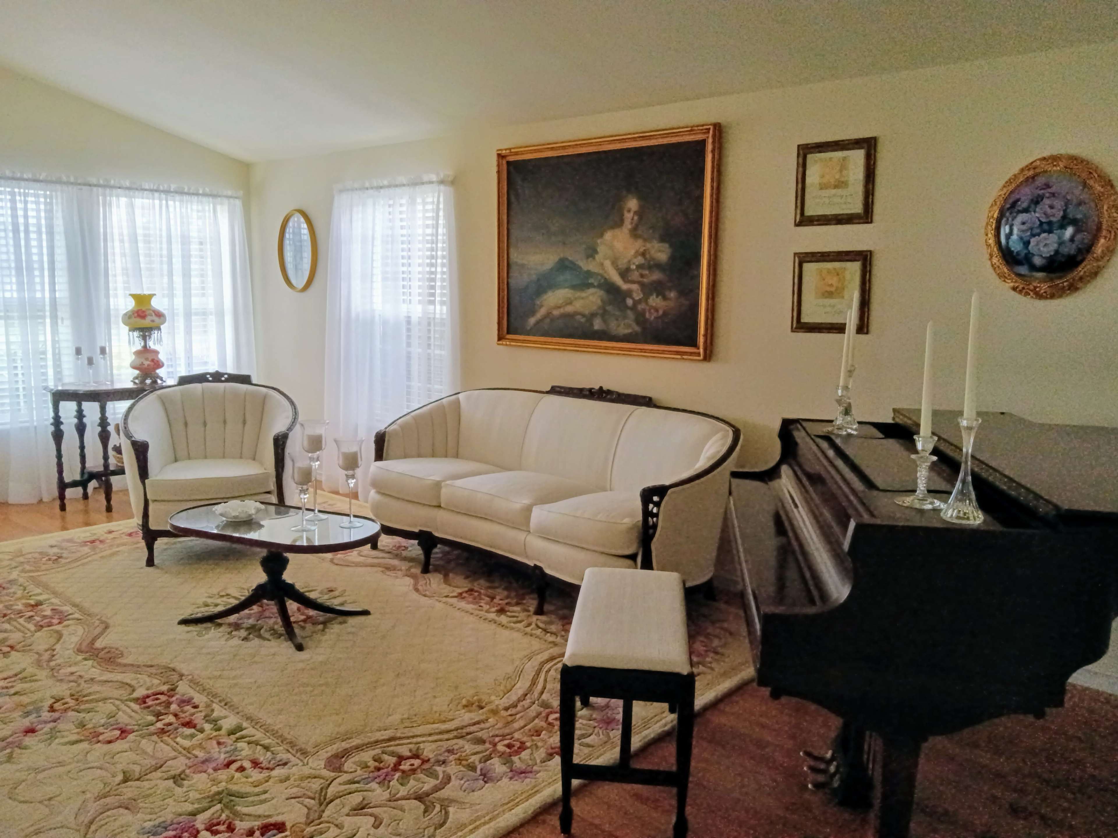 The image shows a light-filled living room with a grand piano, a white sofa, two chairs, a coffee table, and a large painting on the wall.