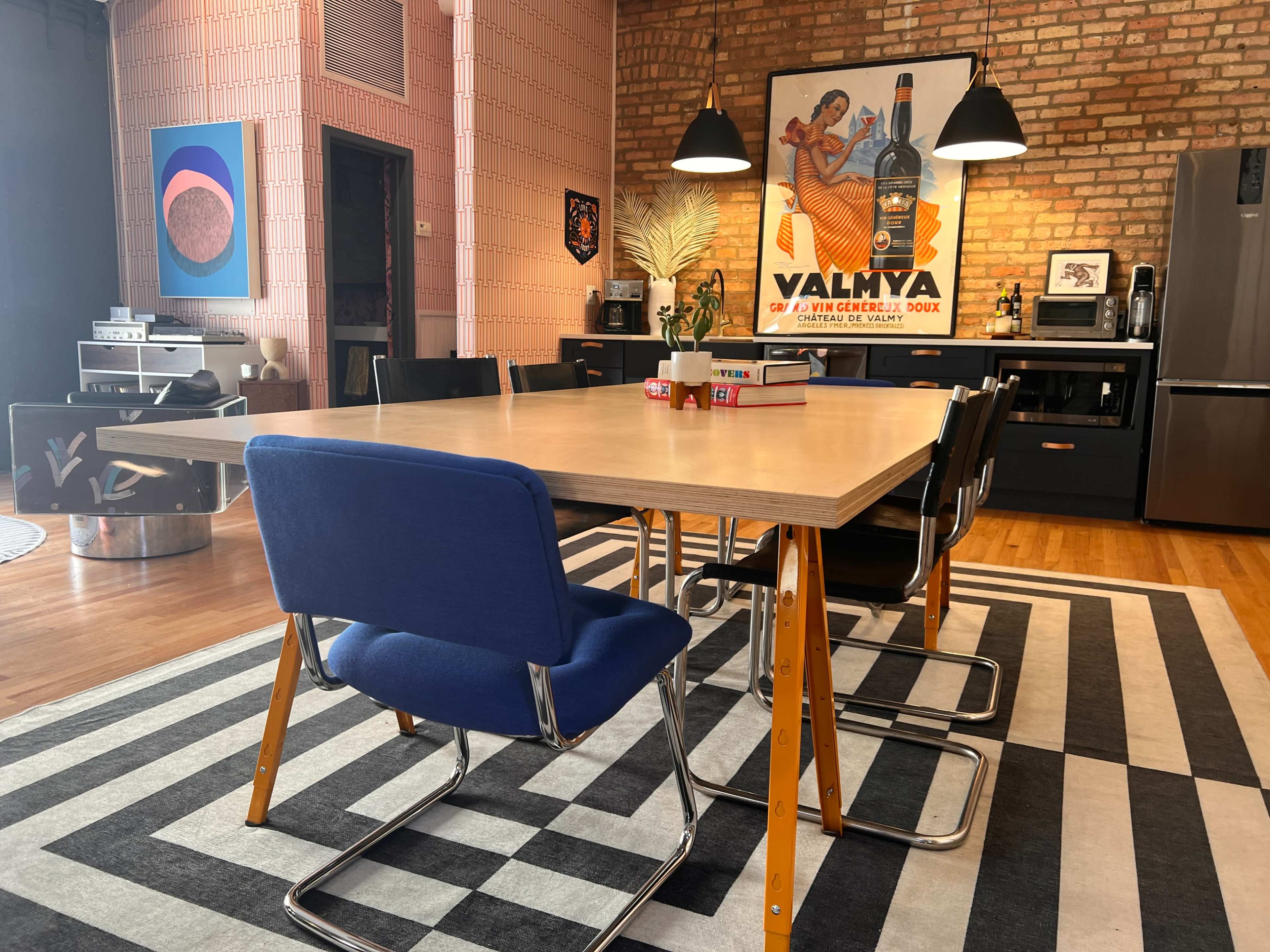 The image depicts a modern dining area featuring a large table surrounded by six chairs on a black and white striped rug, with exposed brick walls and various artworks on display.