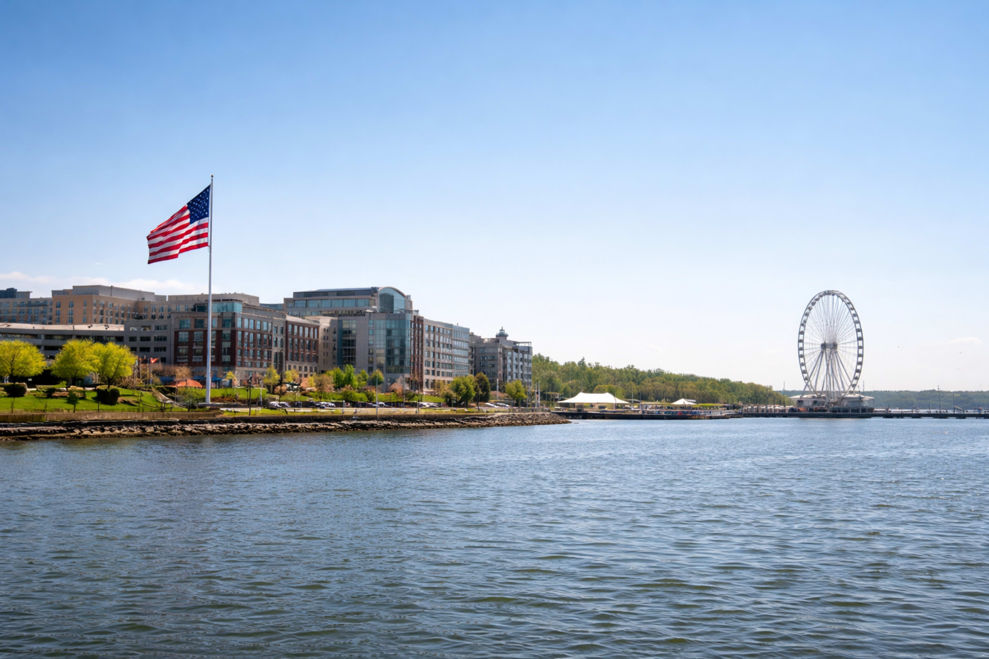 The image shows a lakeside view featuring a large American flag, a riverside building, and a ferris wheel in the distance under clear blue skies.