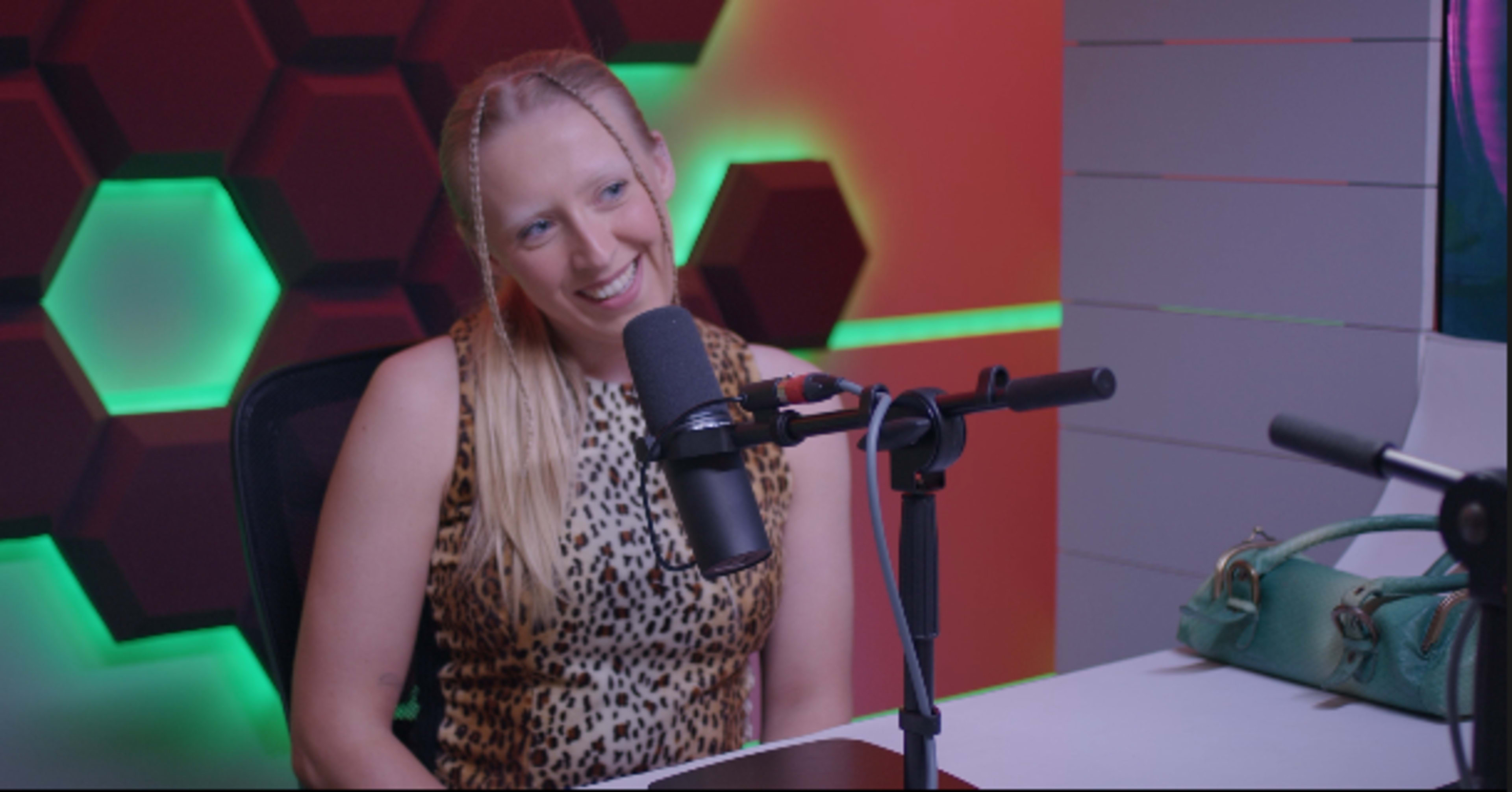 A woman with long hair sits at a table with a microphone, smiling in front of a colorful, patterned backdrop.