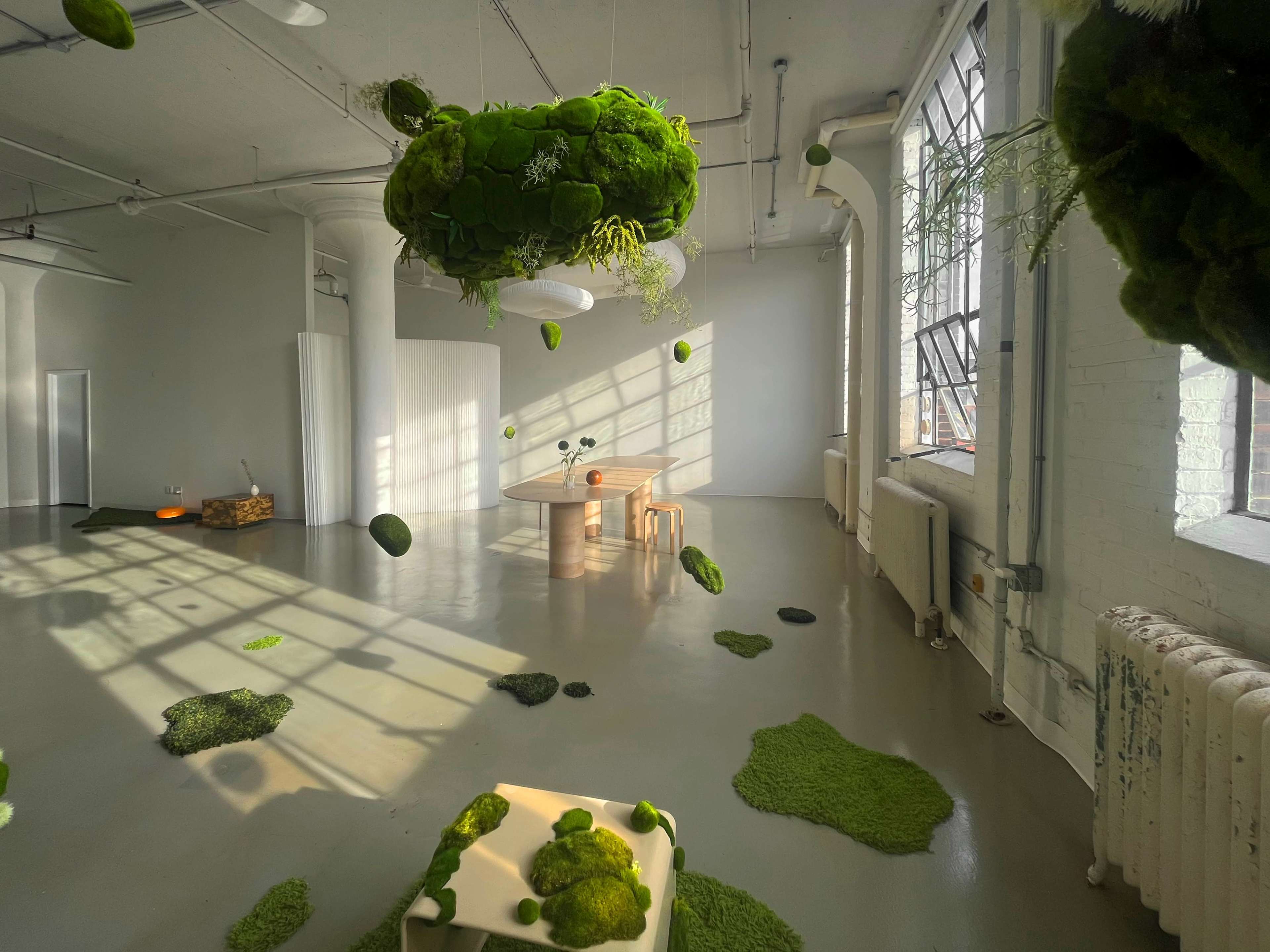 The image shows a spacious, minimalistic room with green moss elements scattered on the floor and a large moss-covered installation hanging from the ceiling.