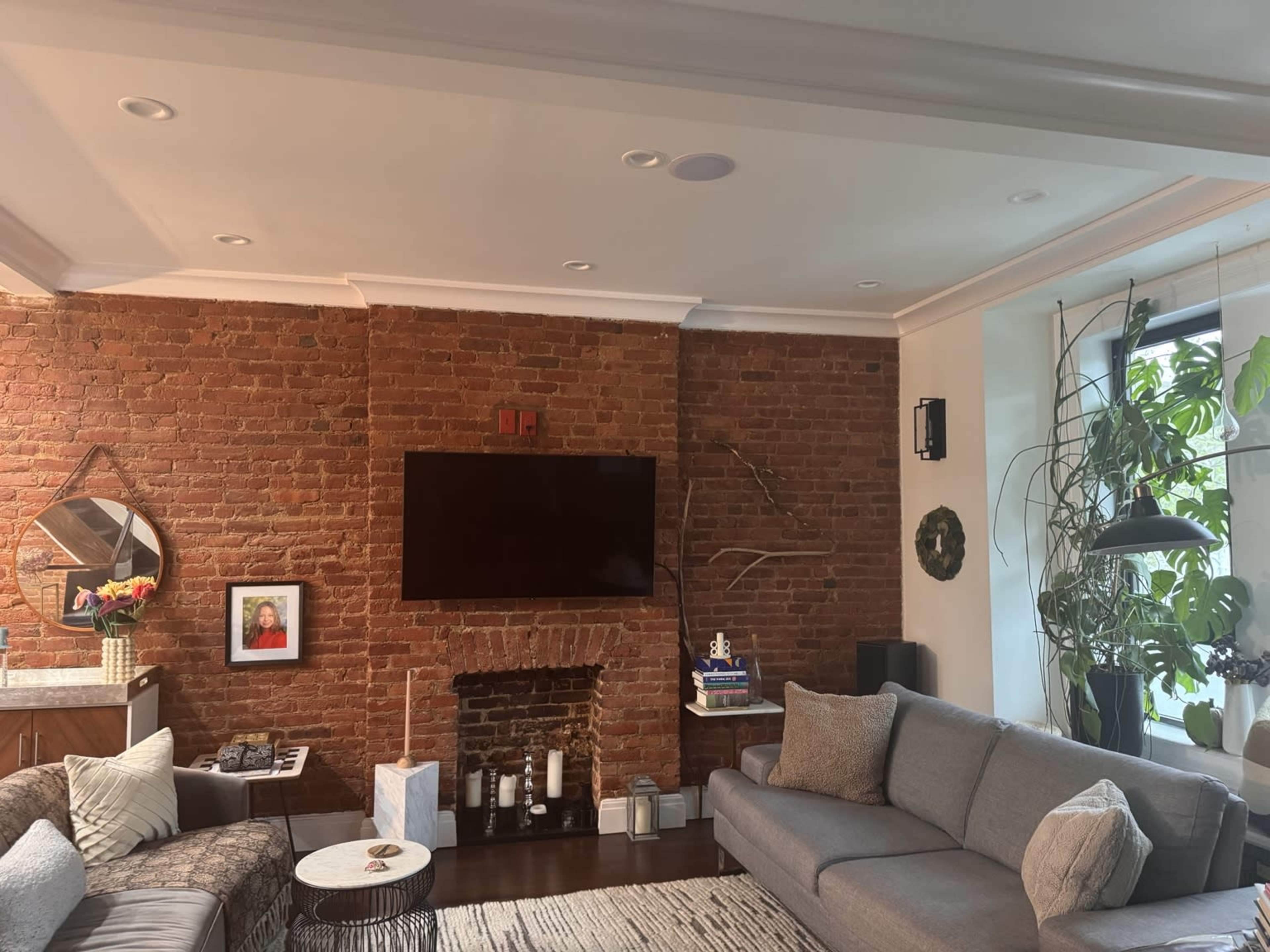 The image shows a living room with a brick wall, a large flat-screen TV mounted above a fireplace, and a sectional sofa arranged across from it.