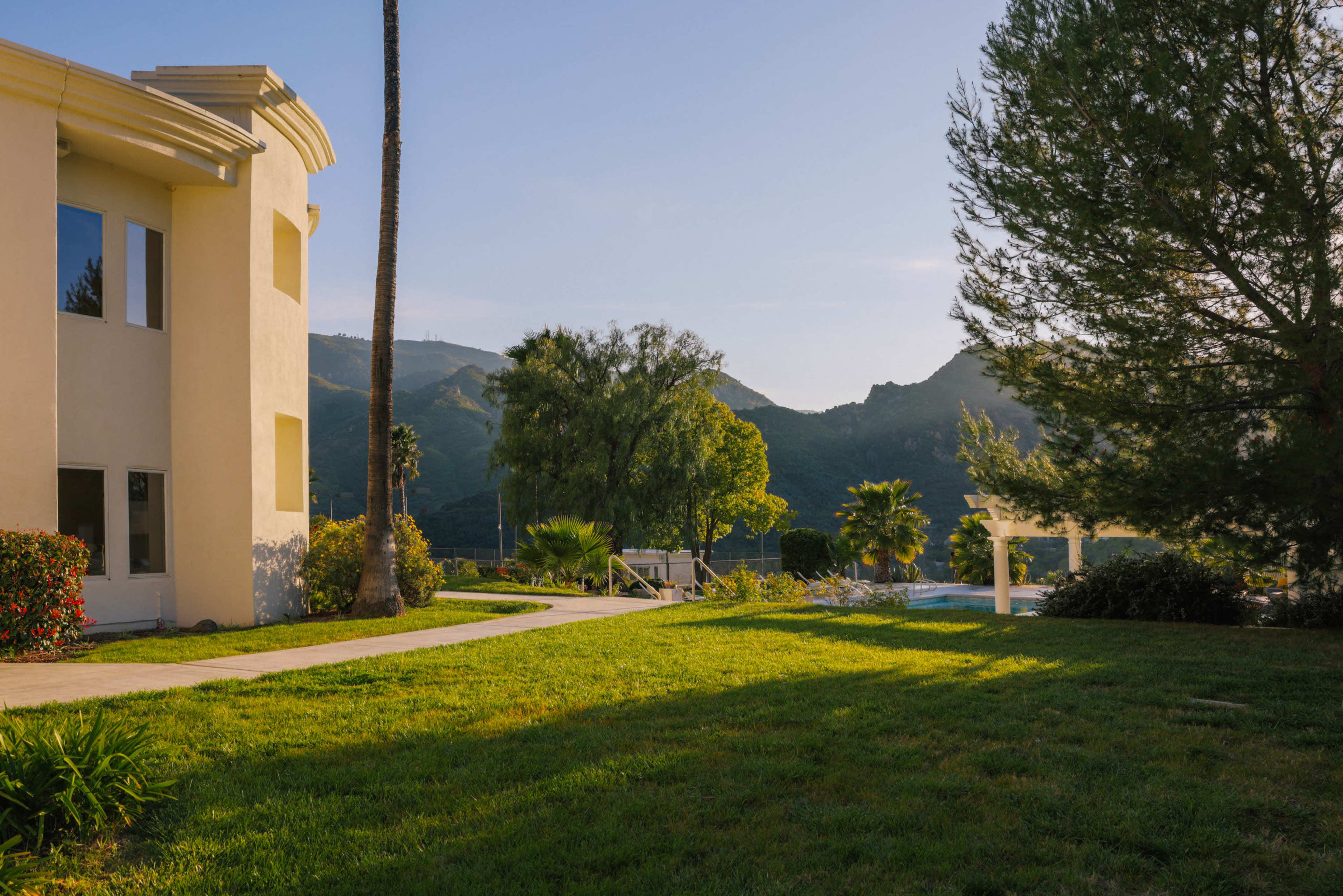 The scene shows a well-manicured lawn next to a modern building, with mountains and palm trees in the background.