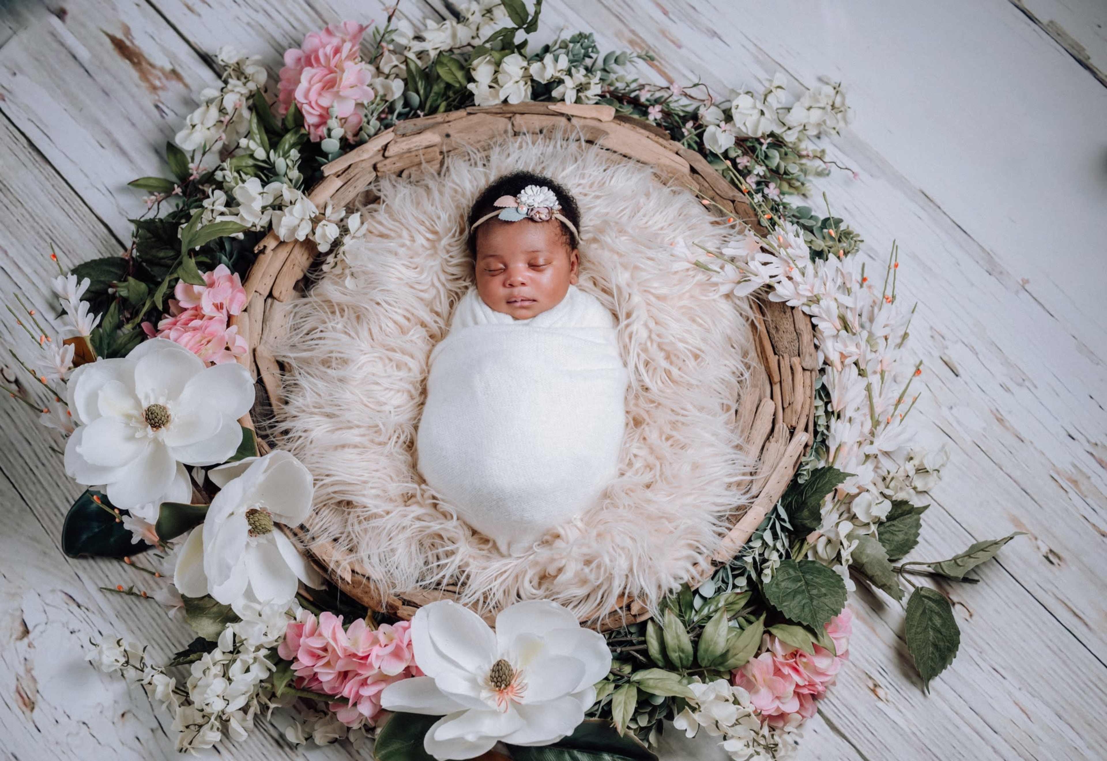 A swaddled baby lies peacefully in a woven basket surrounded by flowers and greenery on a light wooden surface.