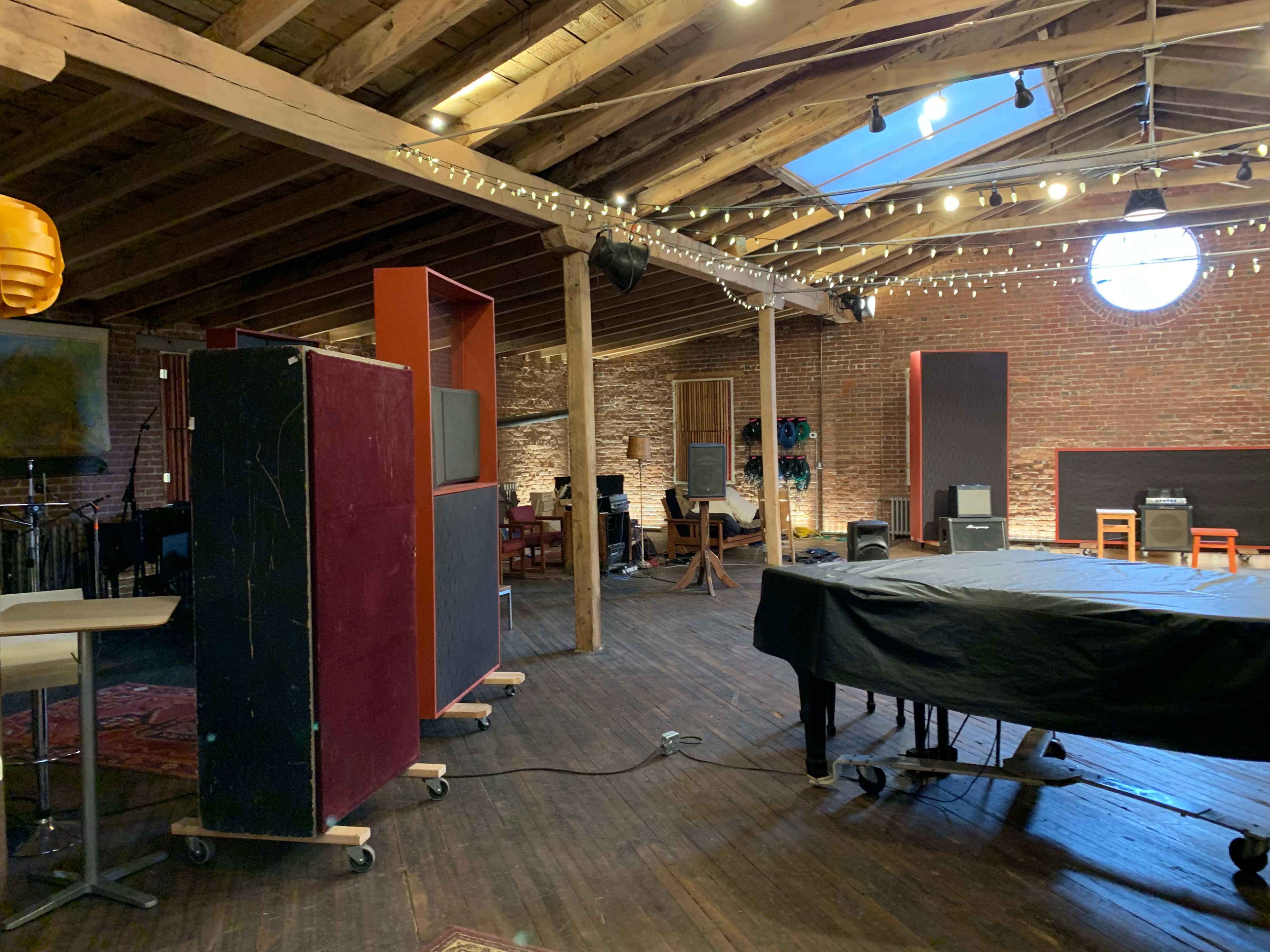 An interior space with wooden beams, brick walls, and scattered furniture including a piano and amplifiers, under soft lighting.