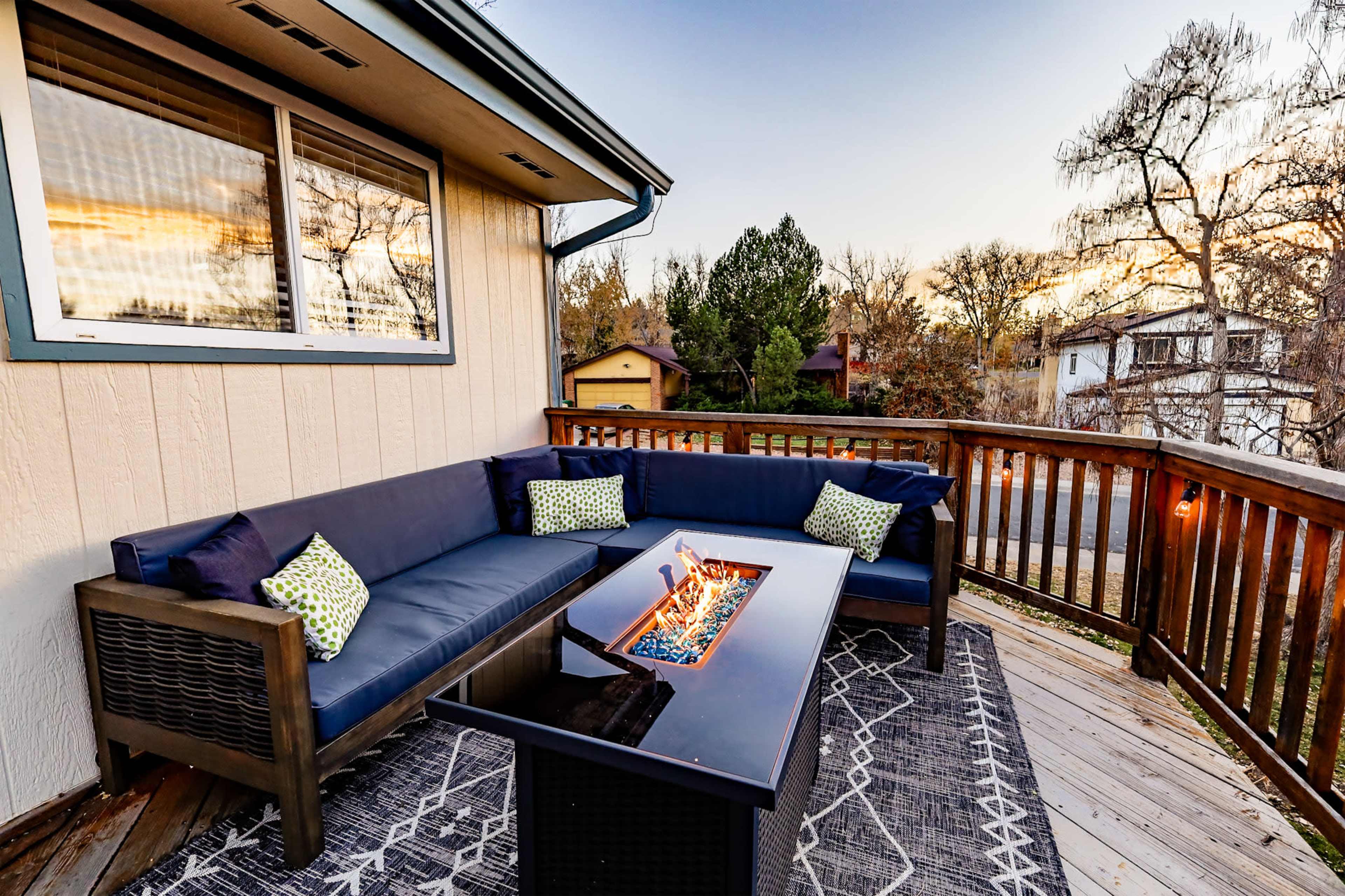 A deck features a sectional sofa with decorative cushions surrounding a fire pit on a wooden surface.