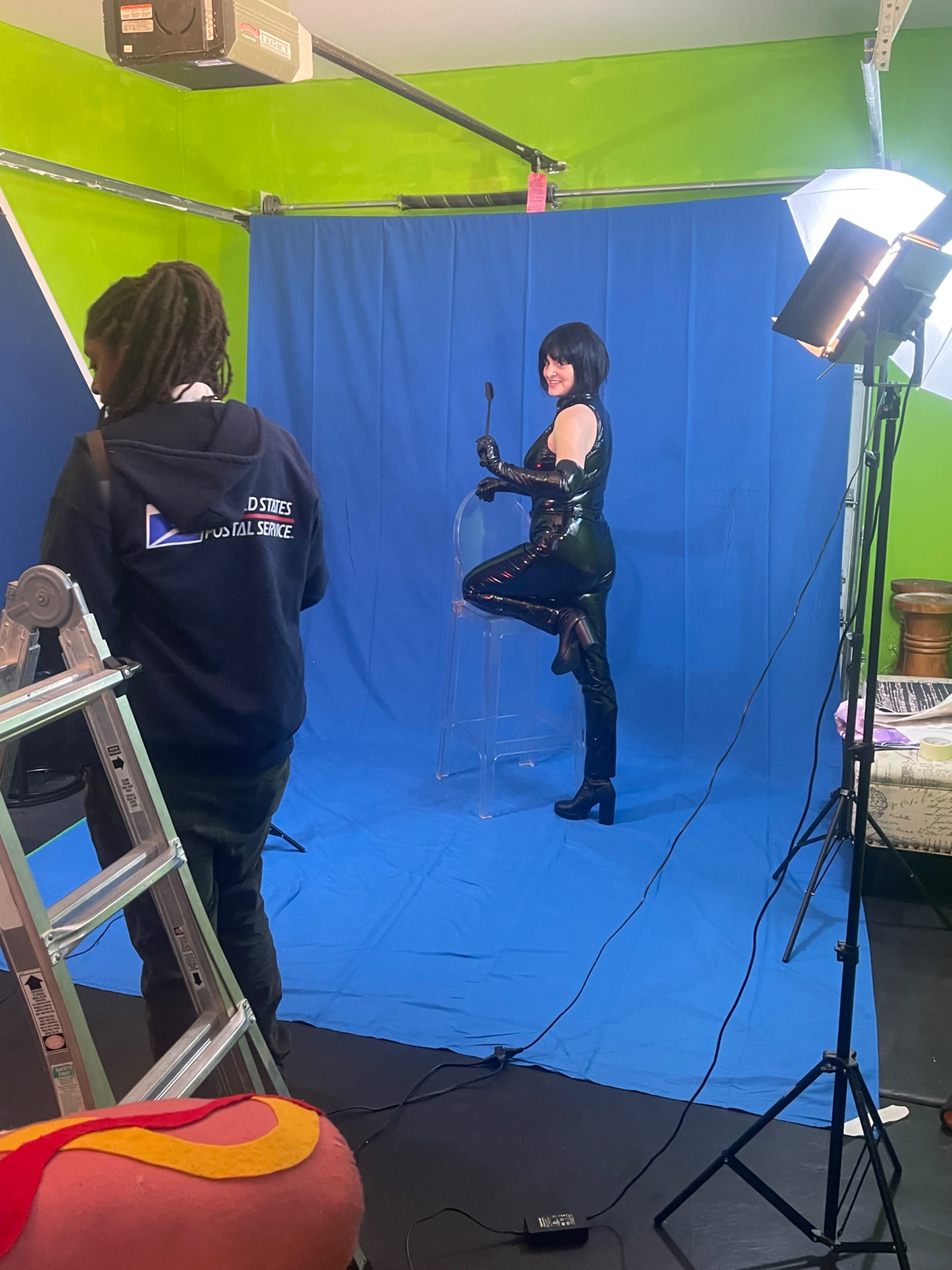 A person in a black outfit poses on a clear chair in front of a blue backdrop while another individual adjusts equipment nearby.