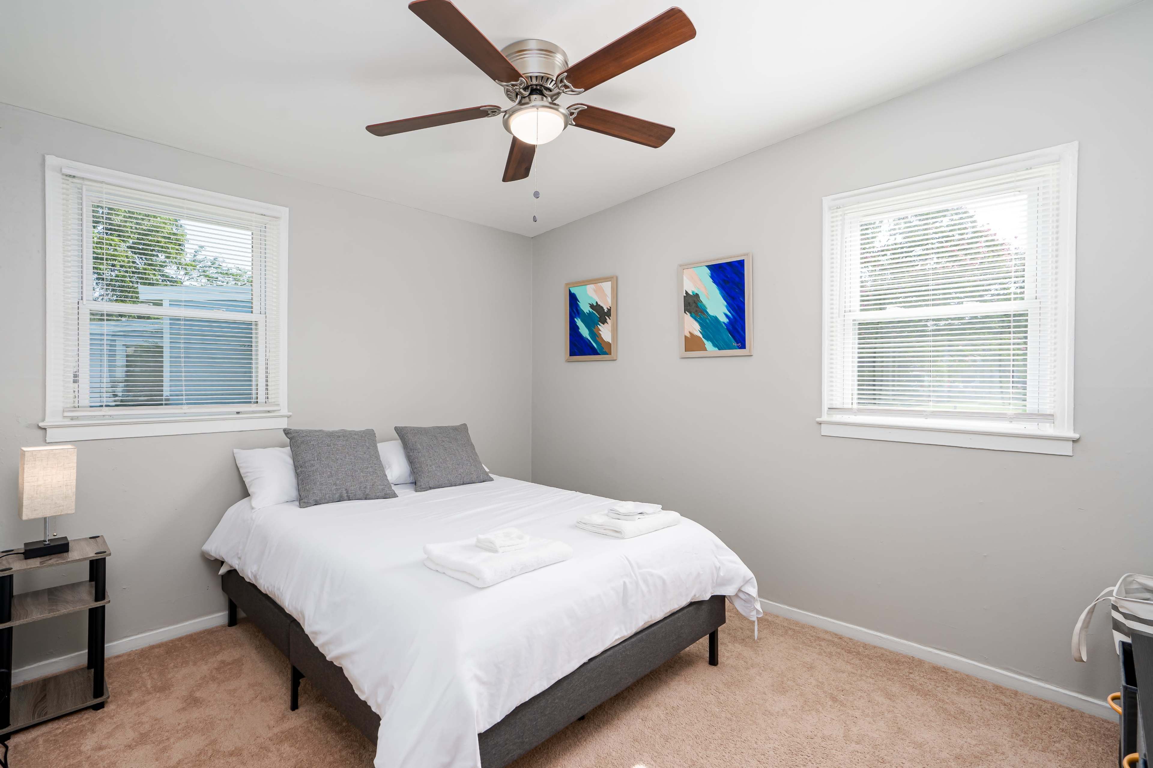A well-lit bedroom features a bed with white linens and two gray pillows, accompanied by a ceiling fan and two framed paintings on the walls.