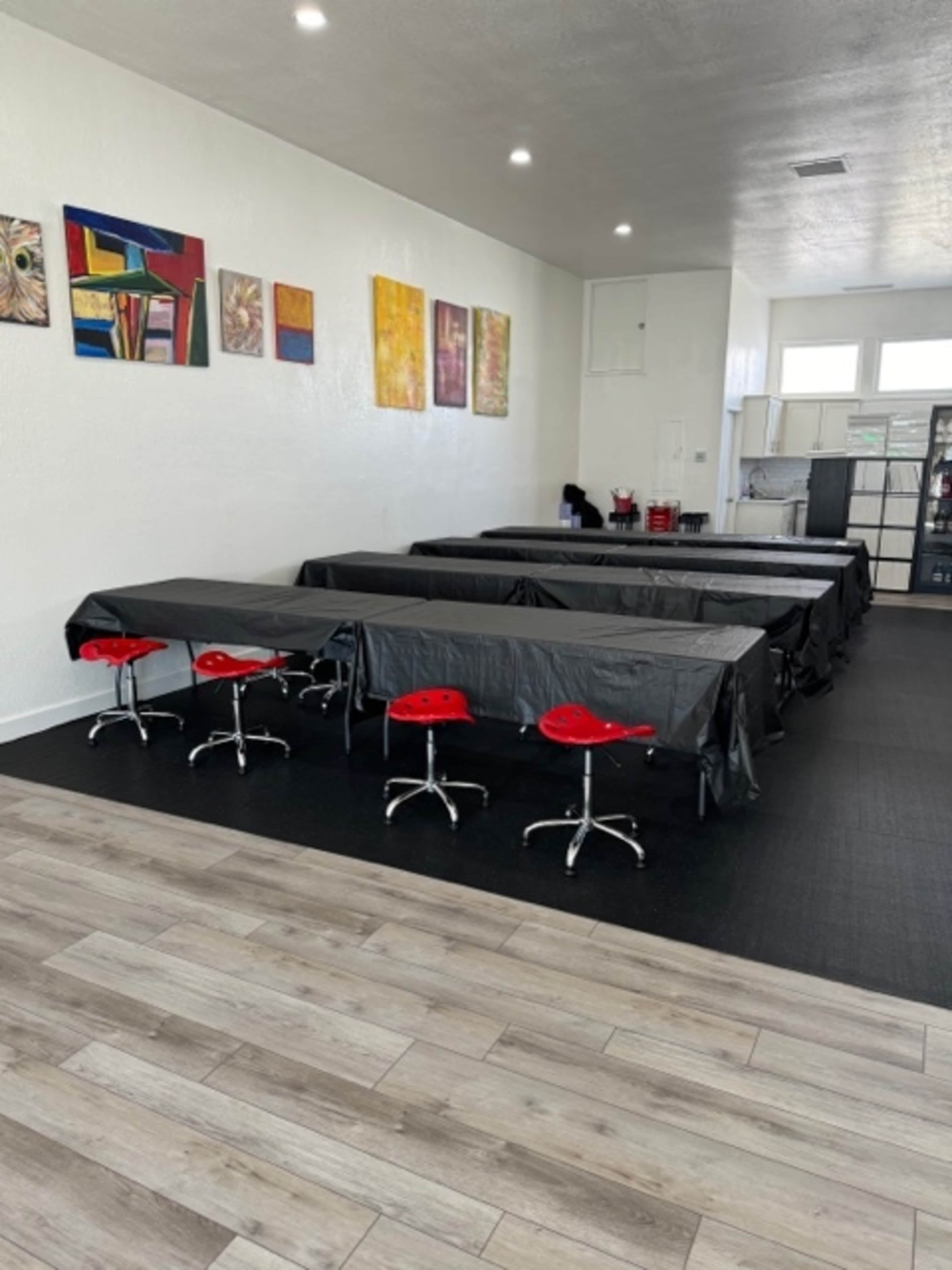A spacious room with multiple black tablecloth-covered tables and red stools, along with colorful paintings on the white walls.