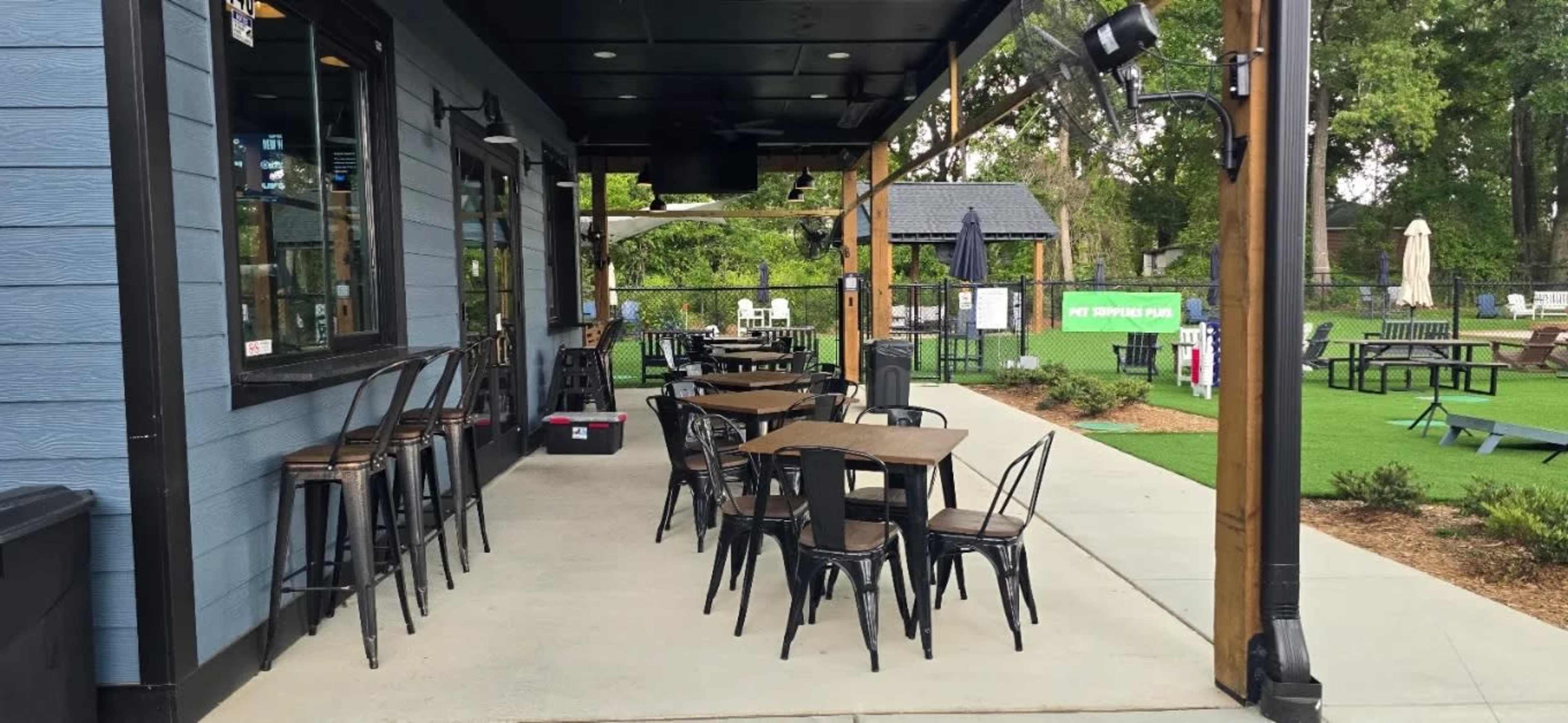The image shows an outdoor dining area with black metal chairs and wooden tables positioned alongside a building, surrounded by a grassy space.