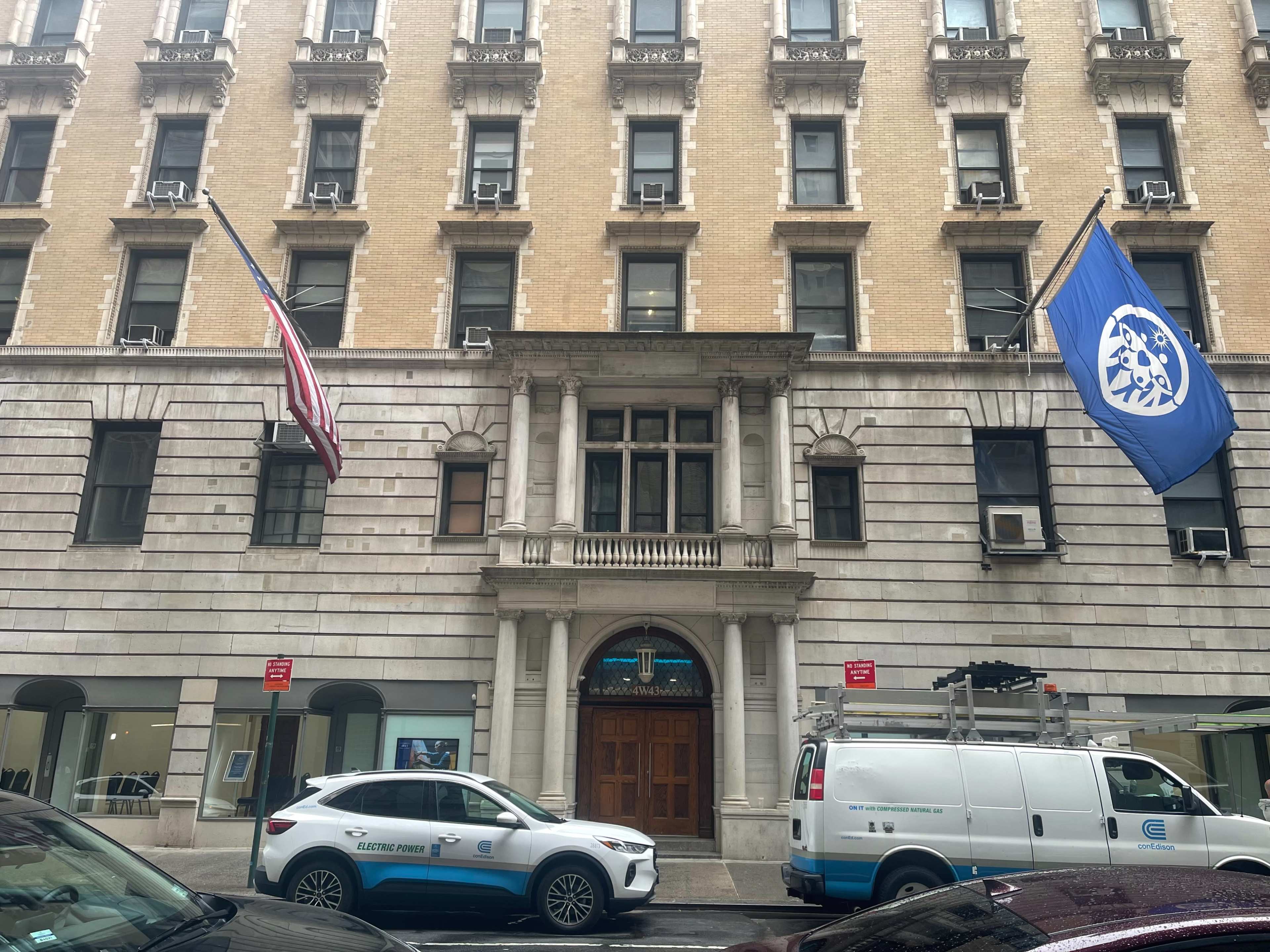 The image shows a building with two flags—an American flag and a blue flag—with a white emblem—mounted above an arched entrance.