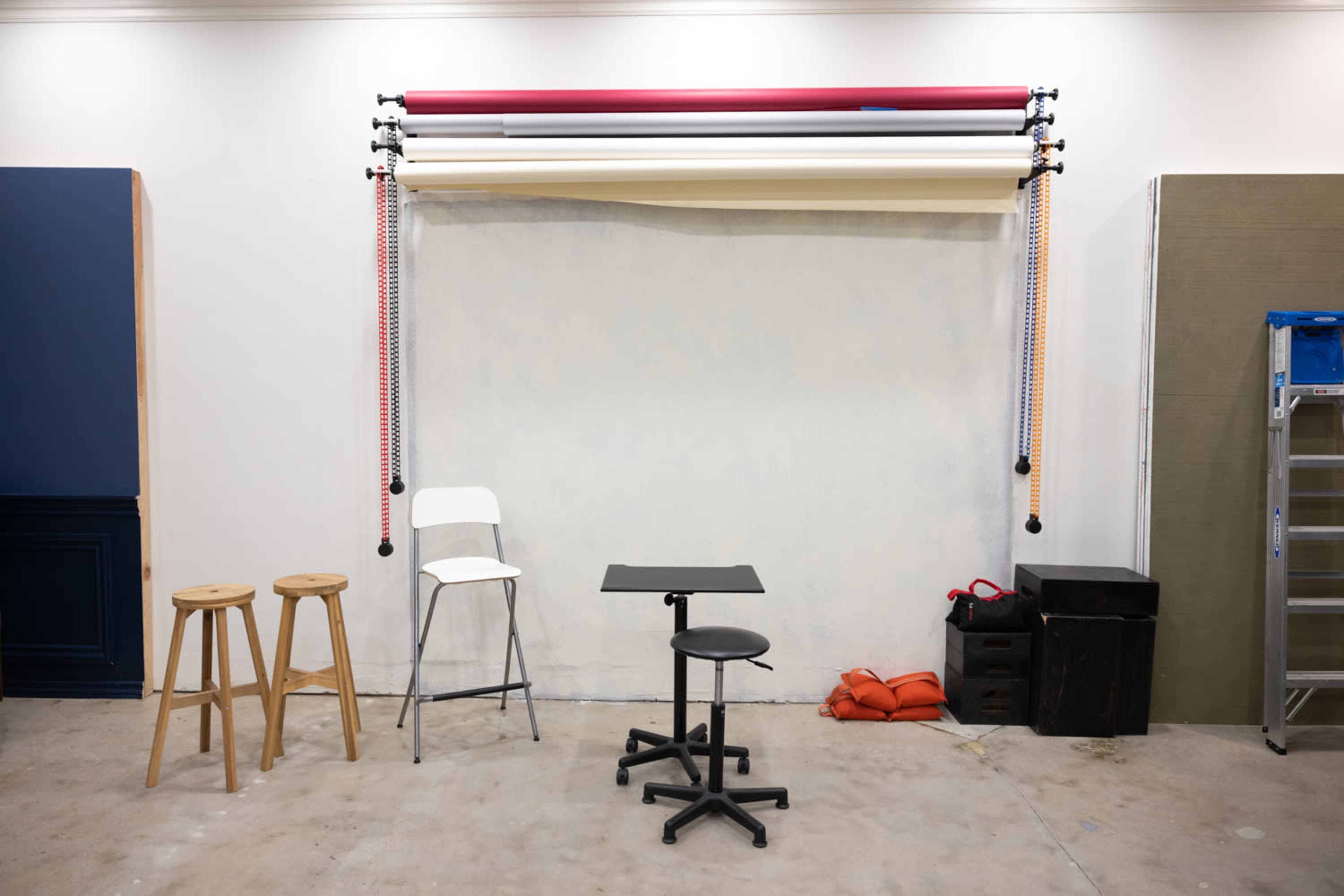 A studio space with a backdrop system featuring rolled paper in various colors, two wooden stools, a white chair, a black table on wheels, and storage bins against the wall.