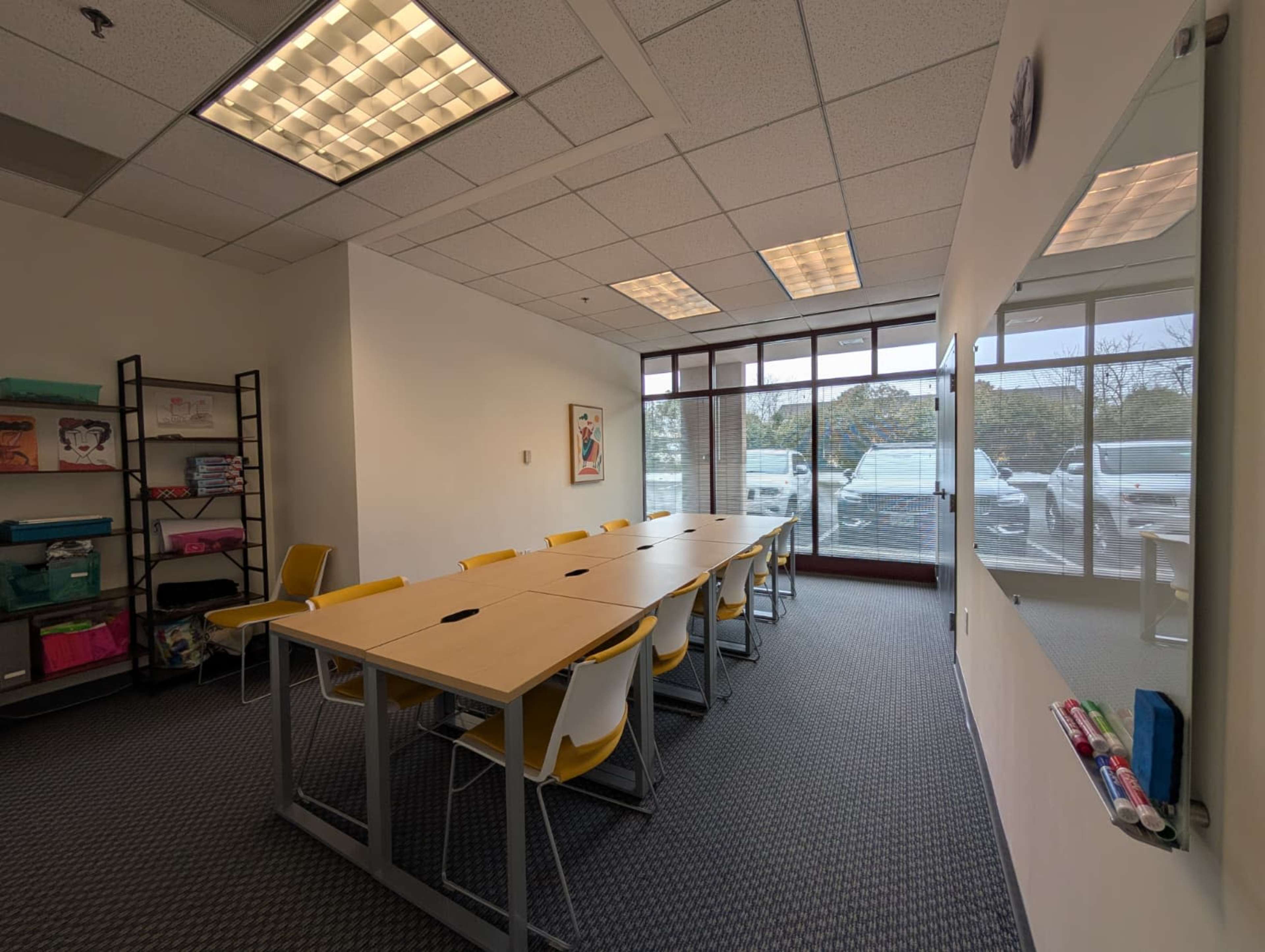 A meeting room features a long table surrounded by yellow chairs, with windows providing natural light from outside.