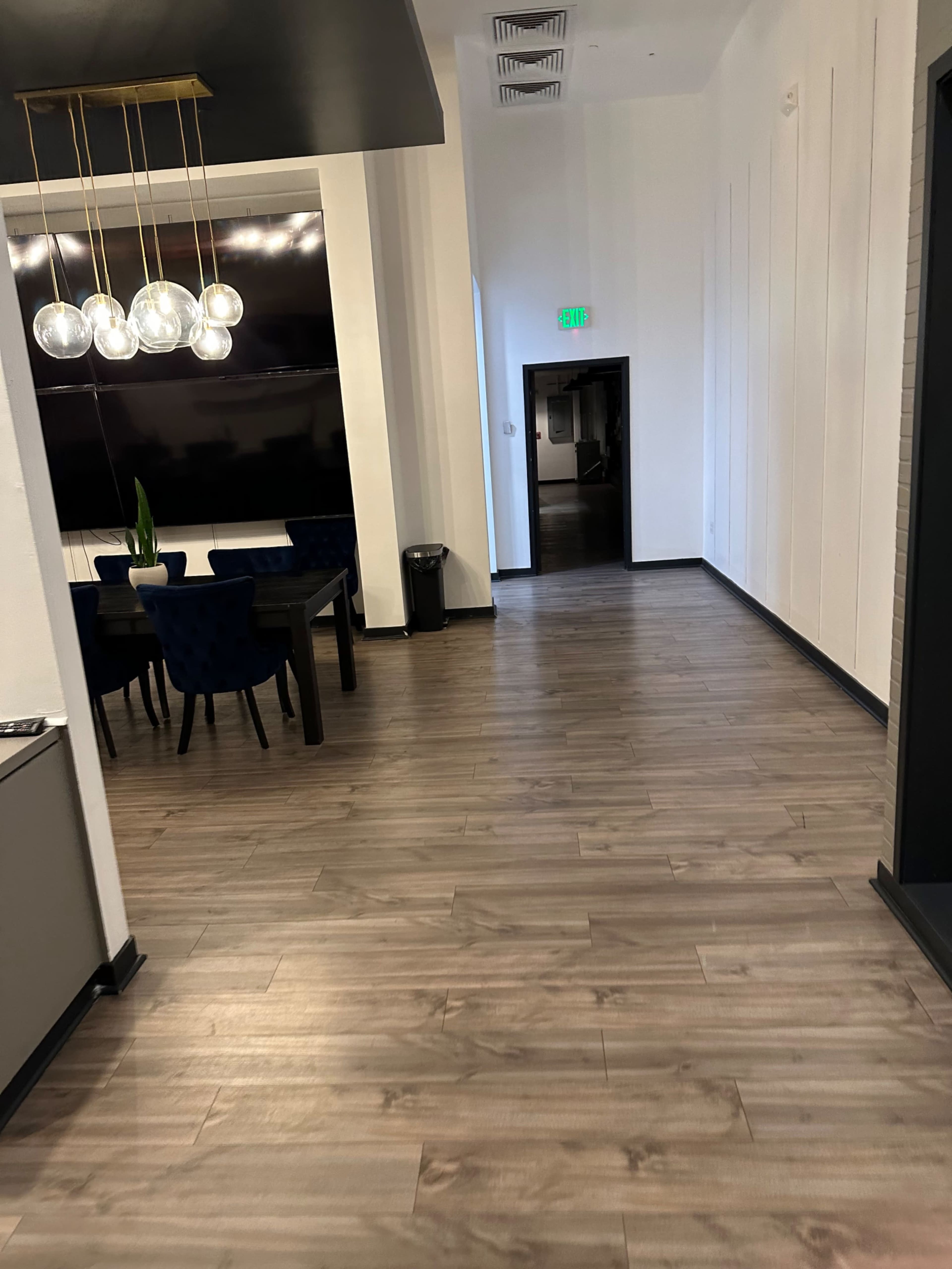 A corridor with wooden flooring leading to a dark doorway, flanked by a dining area on the left with a large table and chairs.