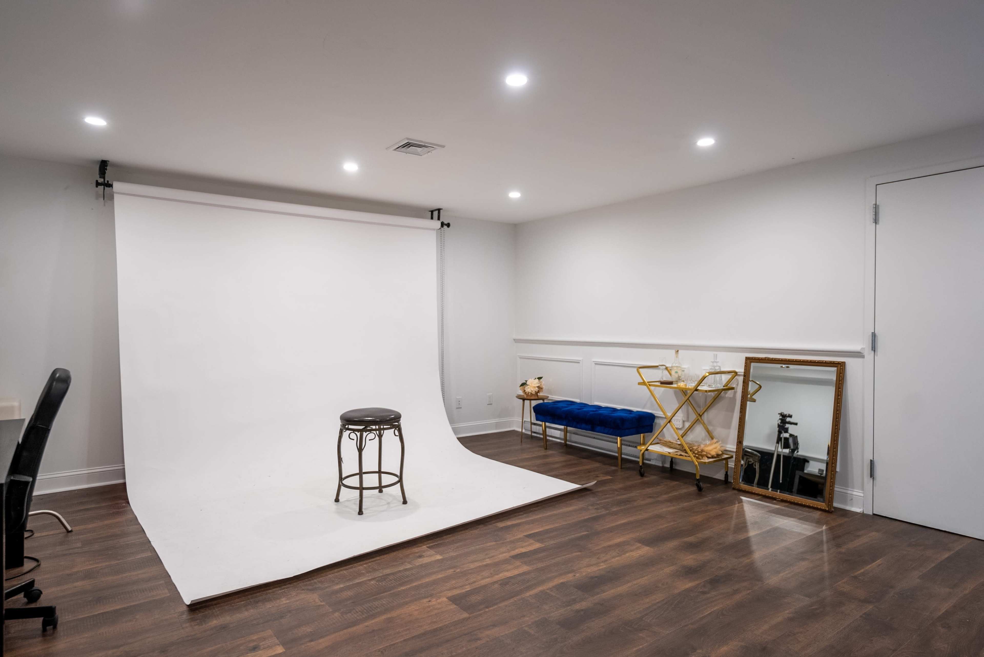 The image depicts a minimalist photo studio featuring a white backdrop, a black stool, a blue bench, and a mirror against the wall.