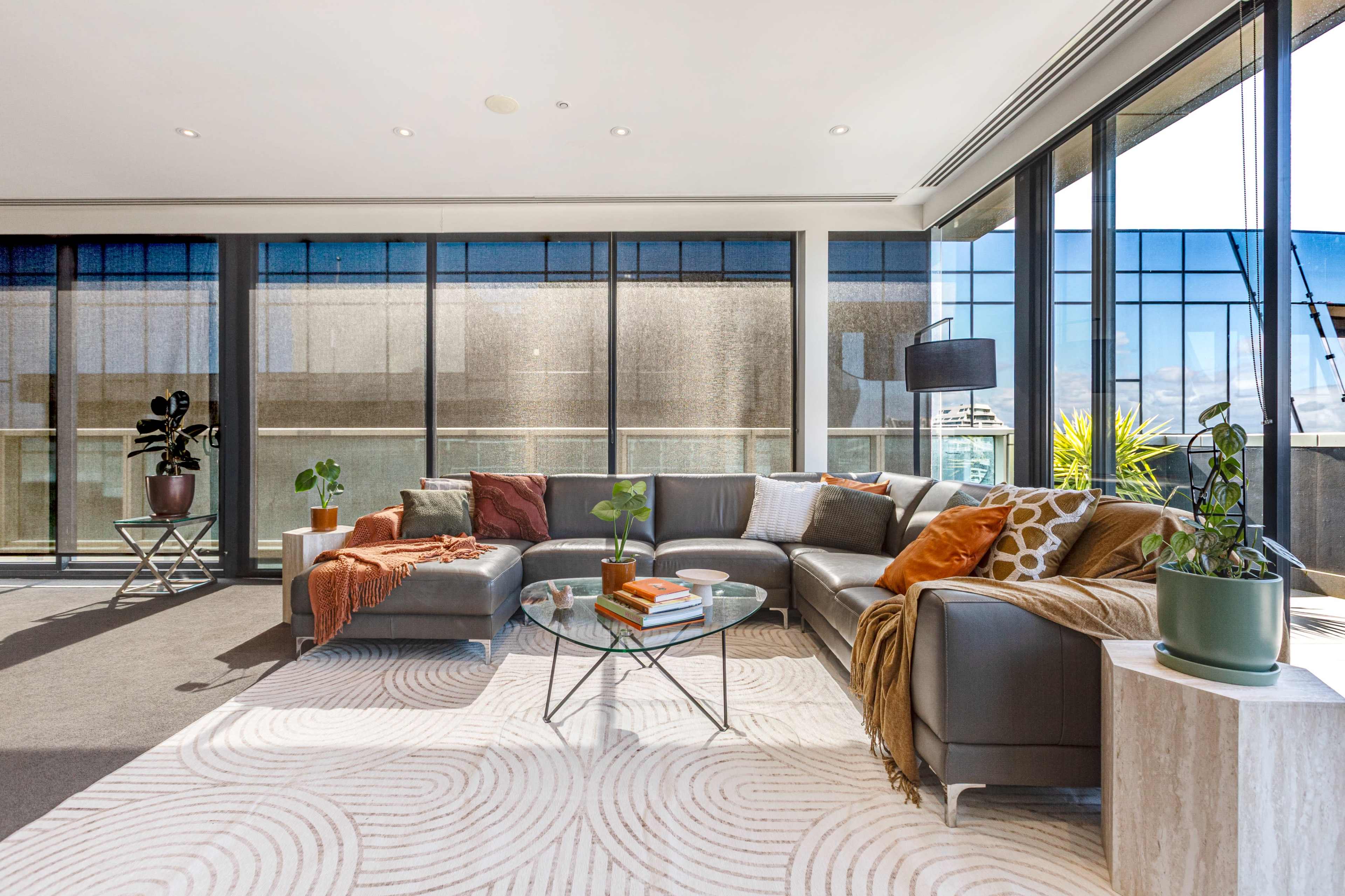 A modern living room features a sectional sofa, glass coffee table, and large windows providing natural light.