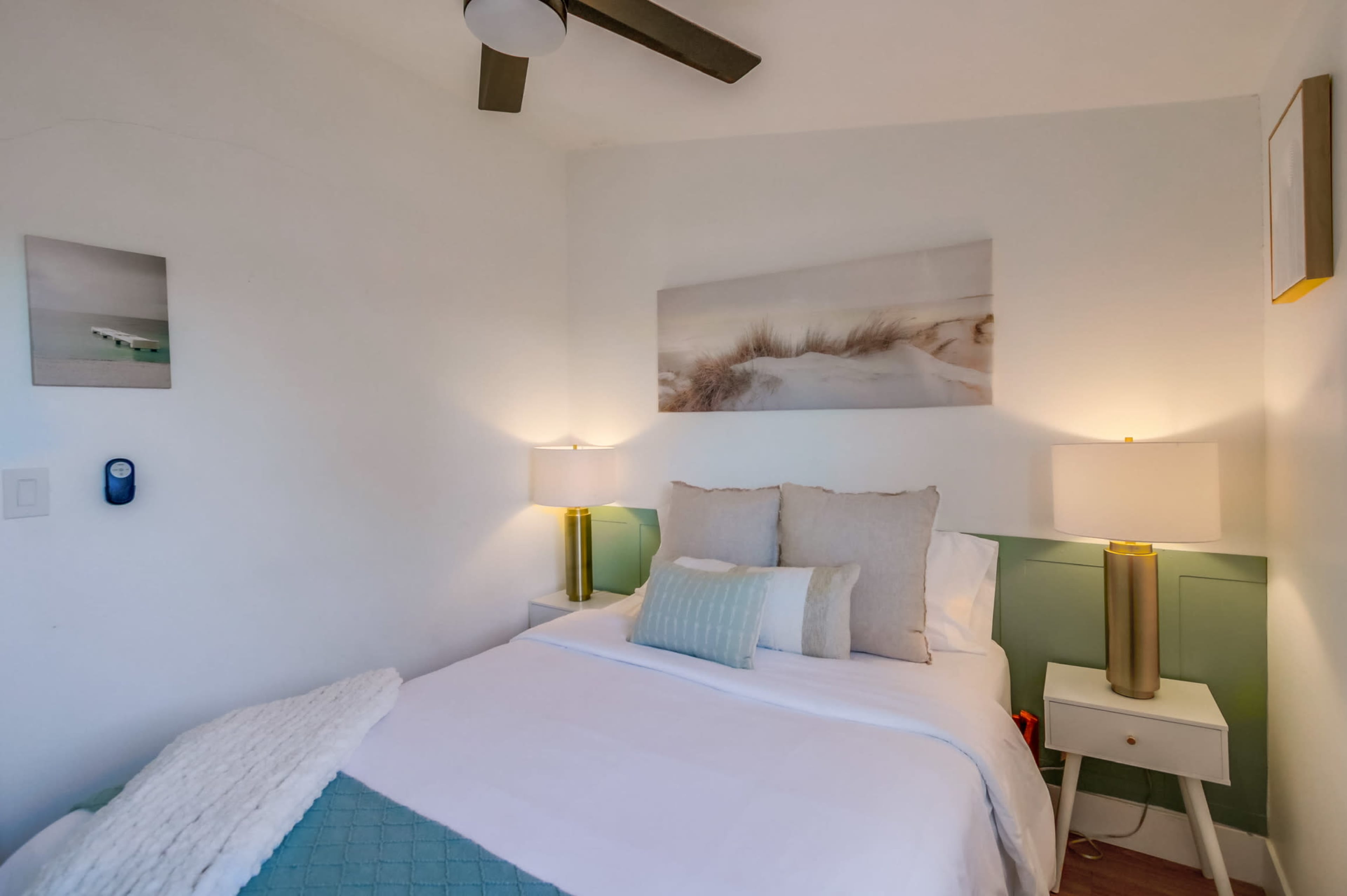 The image shows a small bedroom with a double bed, two bedside lamps, and a coastal-themed artwork above the headboard.