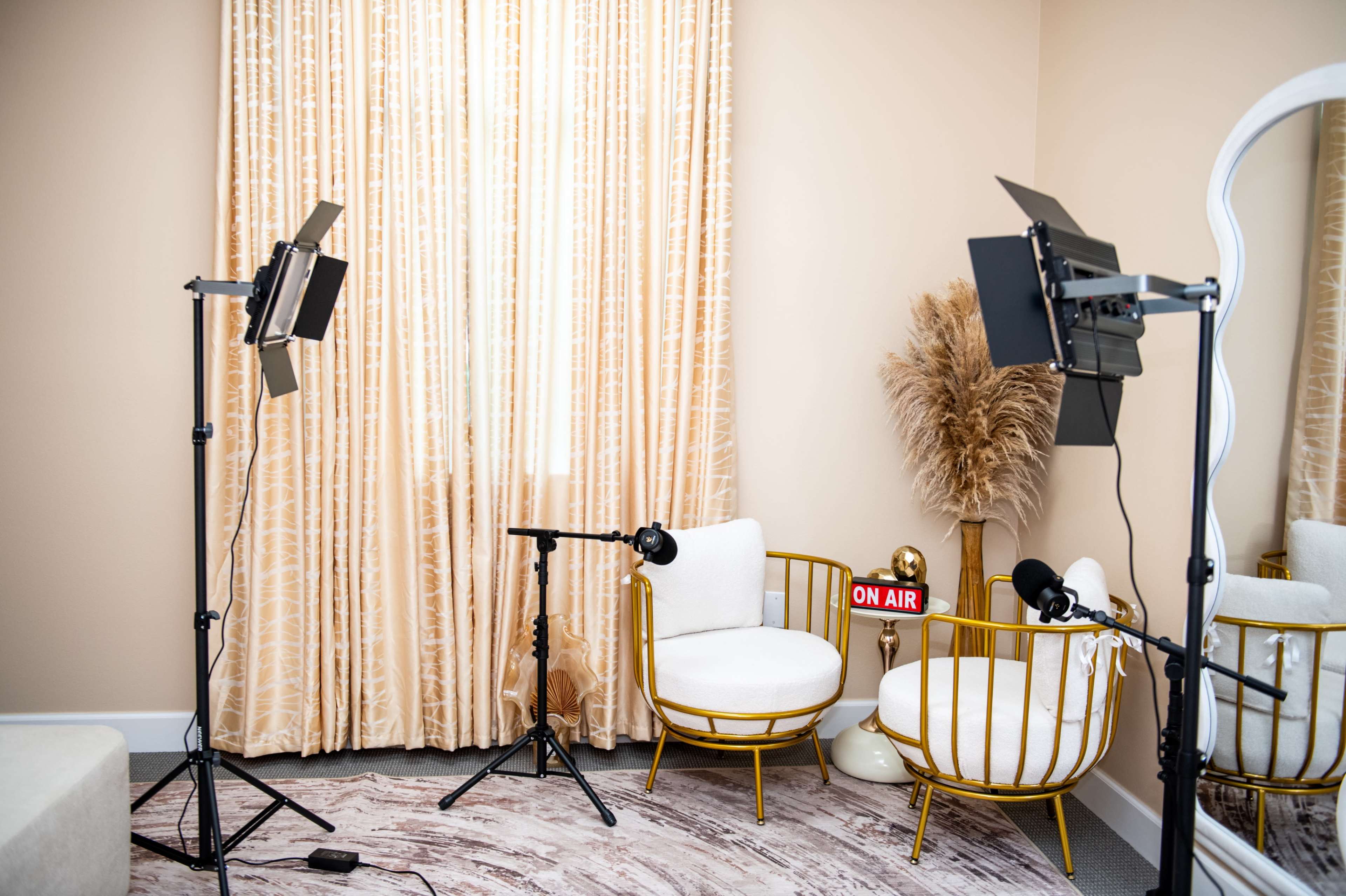 A well-lit room features two gold-colored chairs with white cushions, a large mirror, and soft curtains, along with studio lights and an "ON AIR" sign.