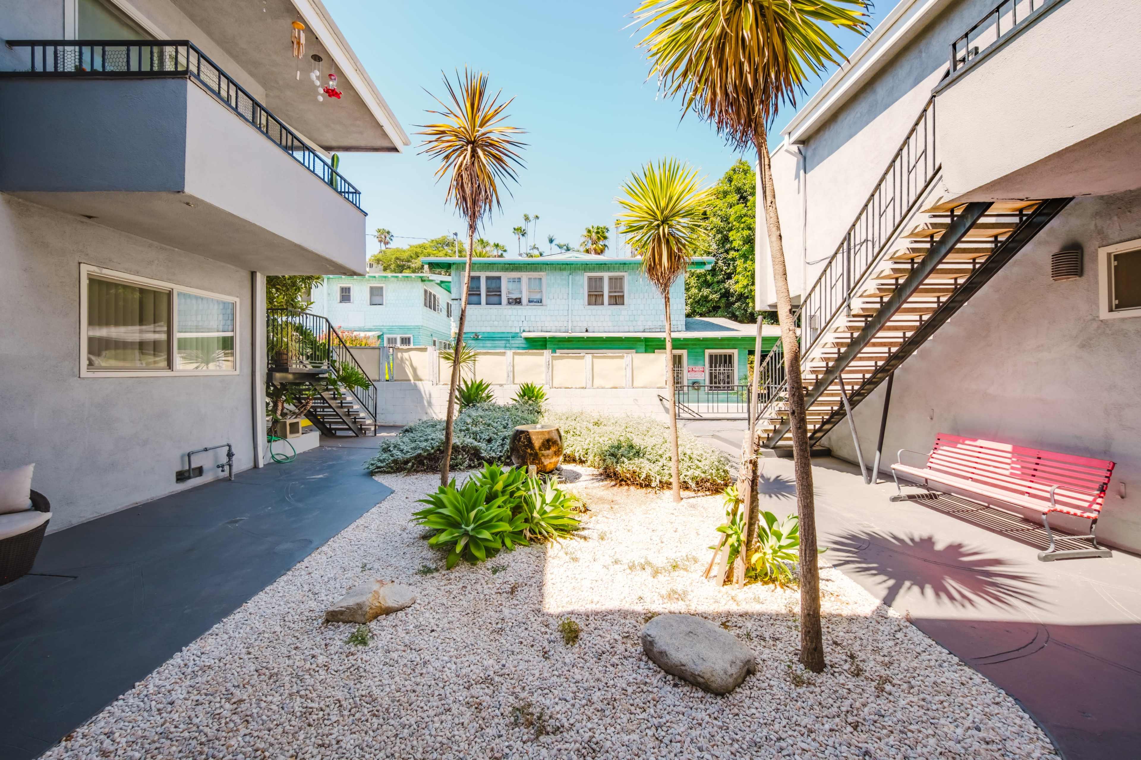 The image shows a landscaped courtyard with palm trees and various plants, adjacent to two multi-story buildings with outdoor staircases.