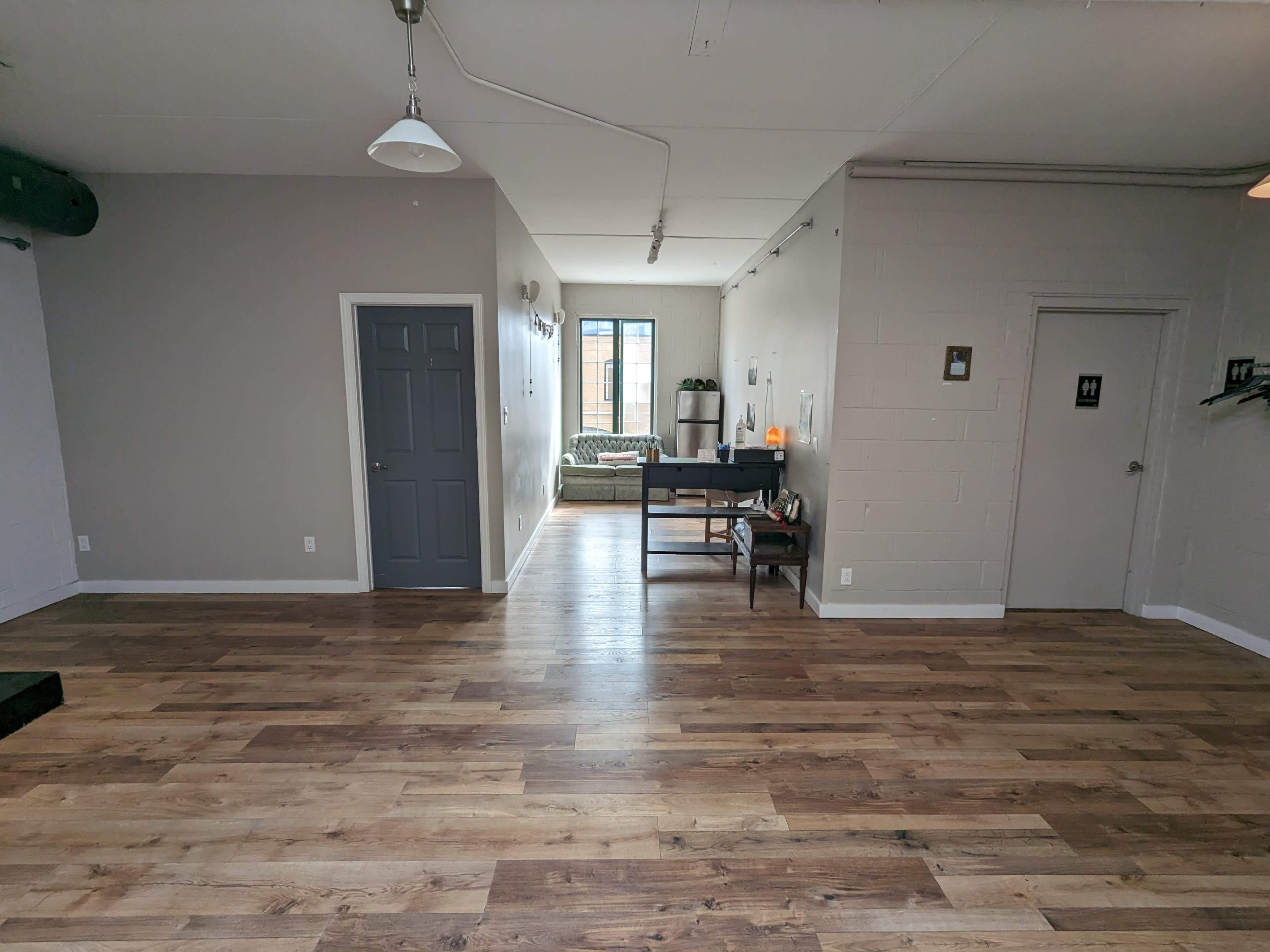 A spacious room with wooden flooring, a gray door on the left, and a table facing a window at the far end.