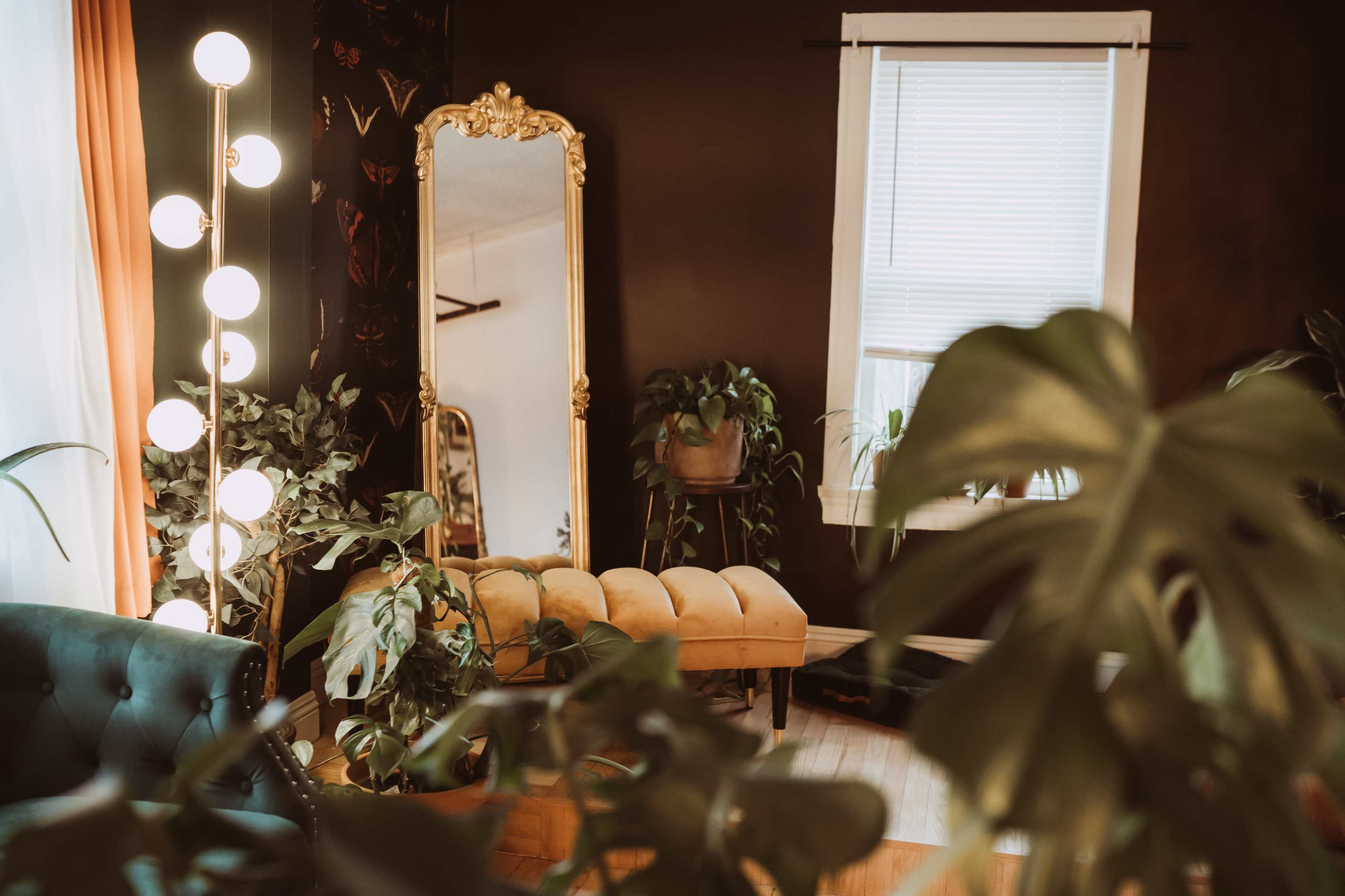 A vintage room features a golden-framed mirror, a yellow tufted bench, and a tall lamp with round bulbs, surrounded by various indoor plants.