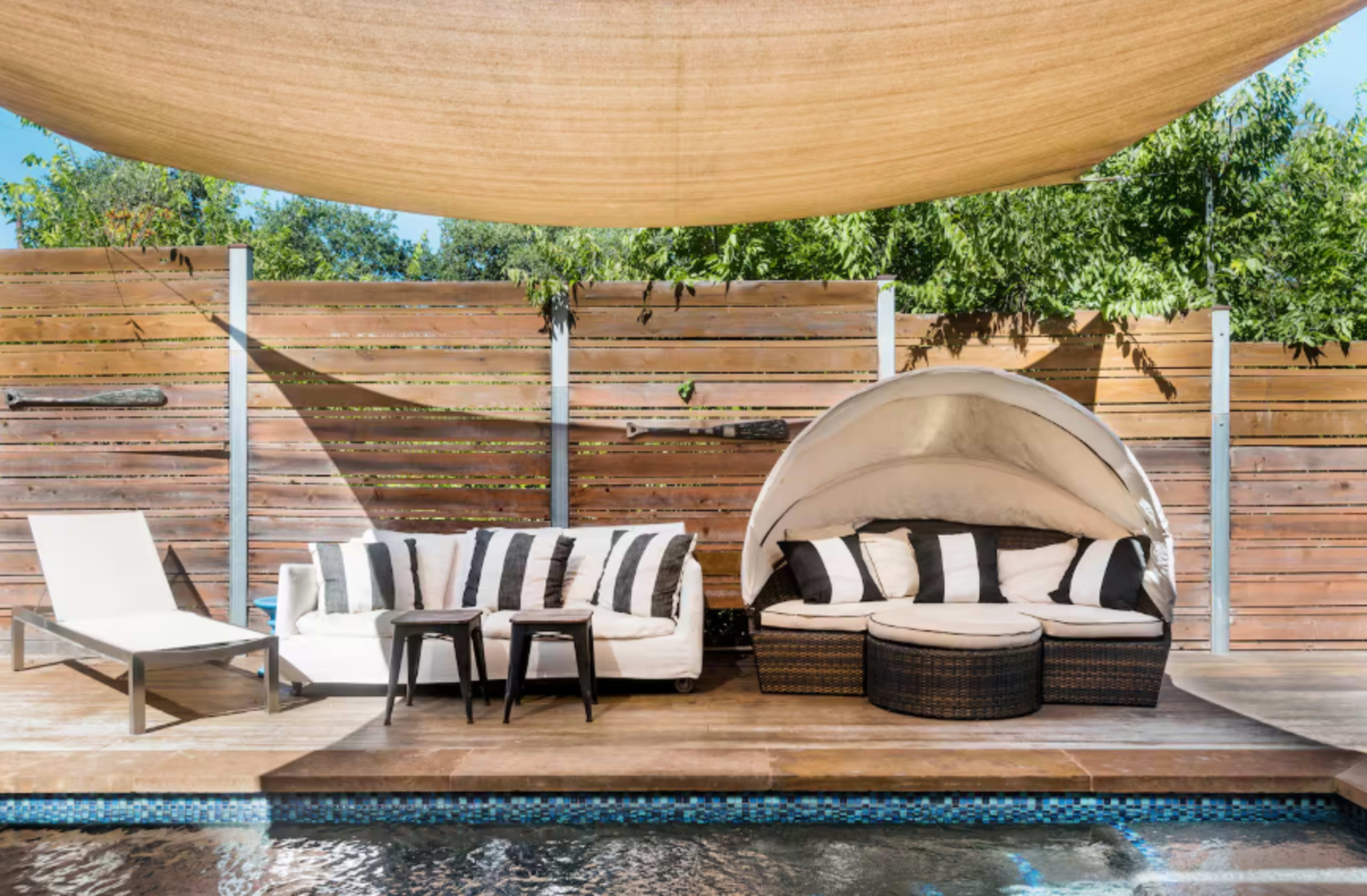 The image shows a stylish outdoor seating area by a pool, featuring a white sofa with black stripes, two small tables, and a round canopy chair under a shaded area.