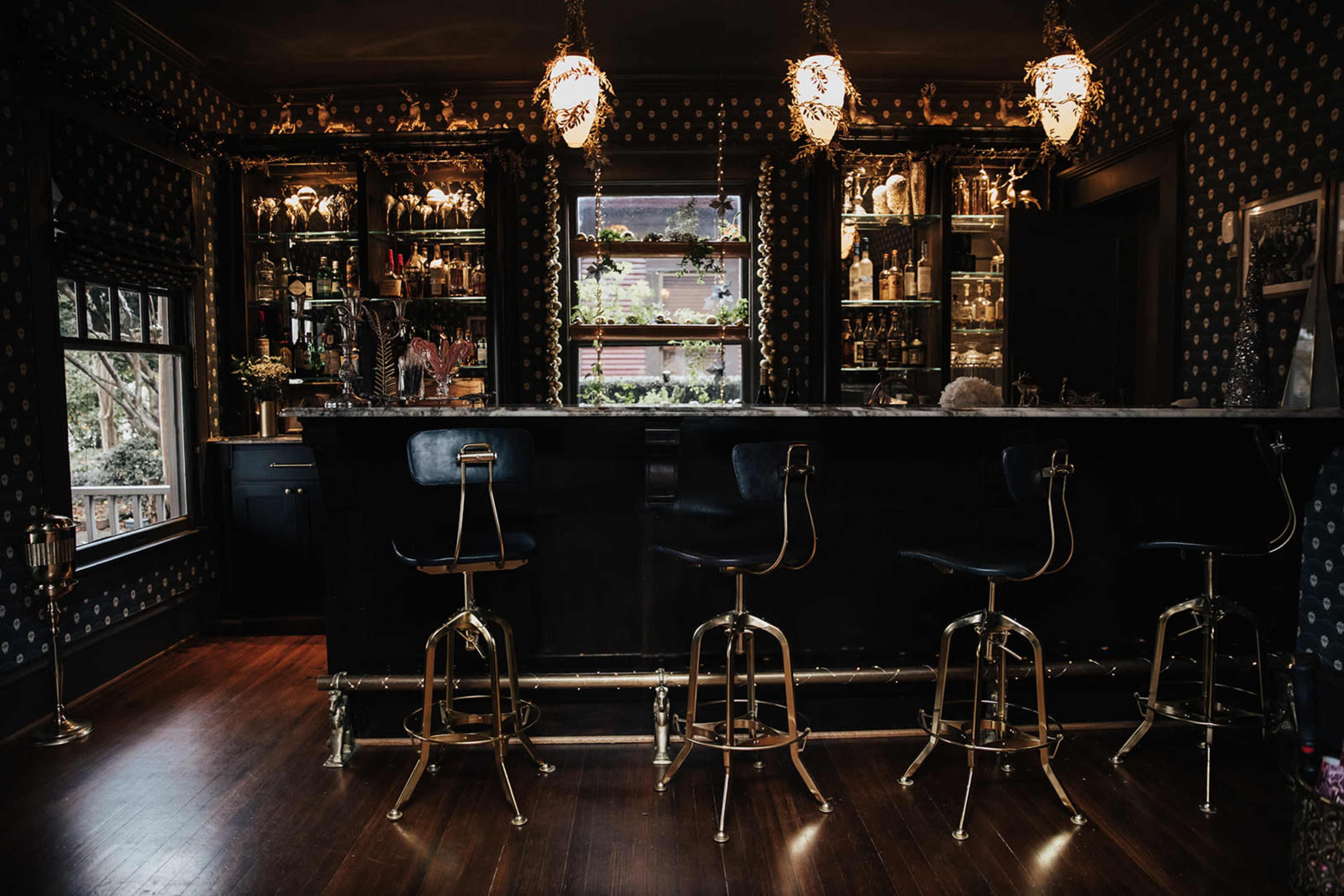 A dimly lit bar area with a dark countertop, gold accents, and several high stools, surrounded by shelves filled with glassware and decor.
