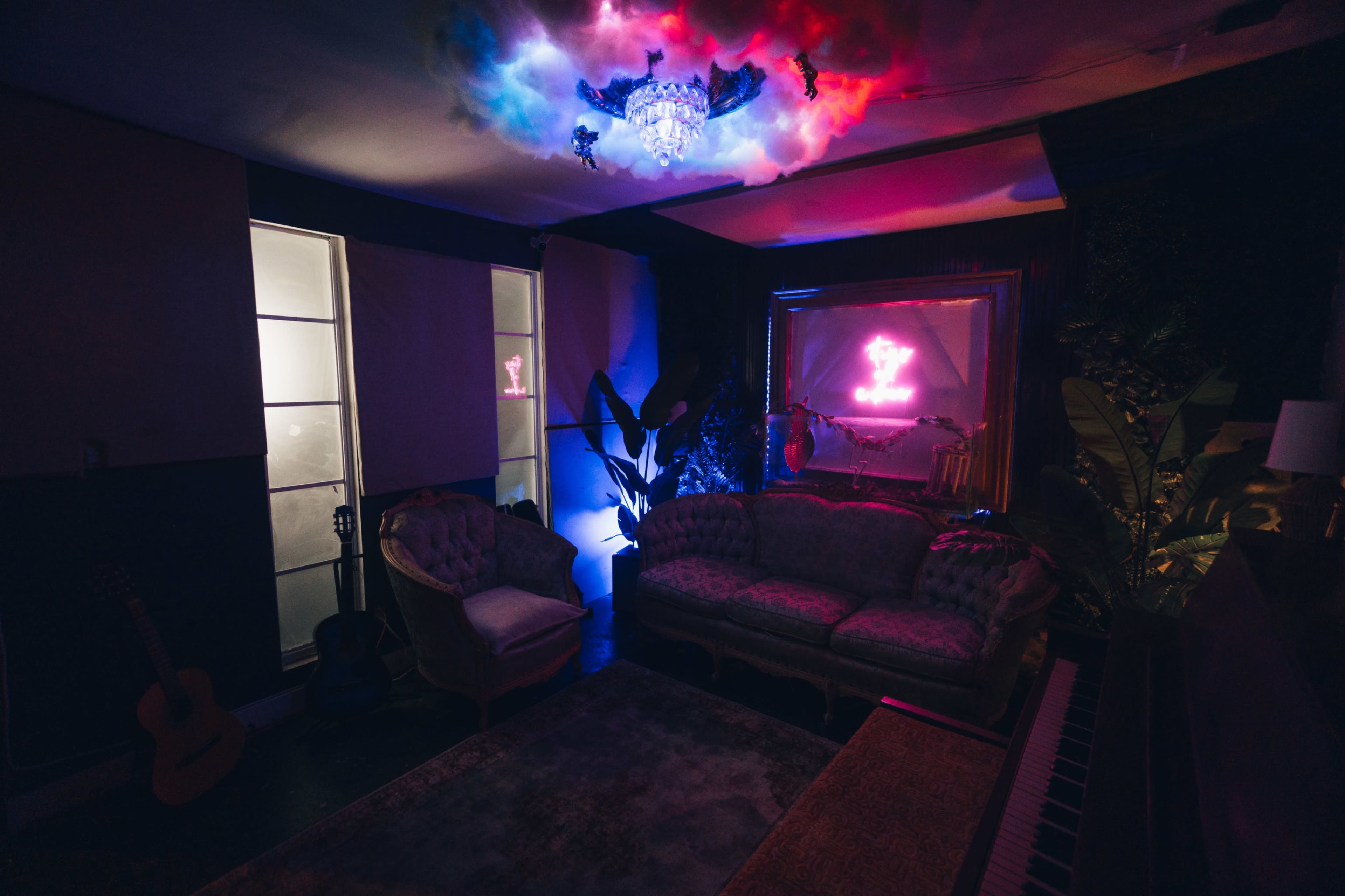 The scene features a dimly lit room with a plush sofa, plants, a guitar, and a chandelier casting colorful lights on the walls.