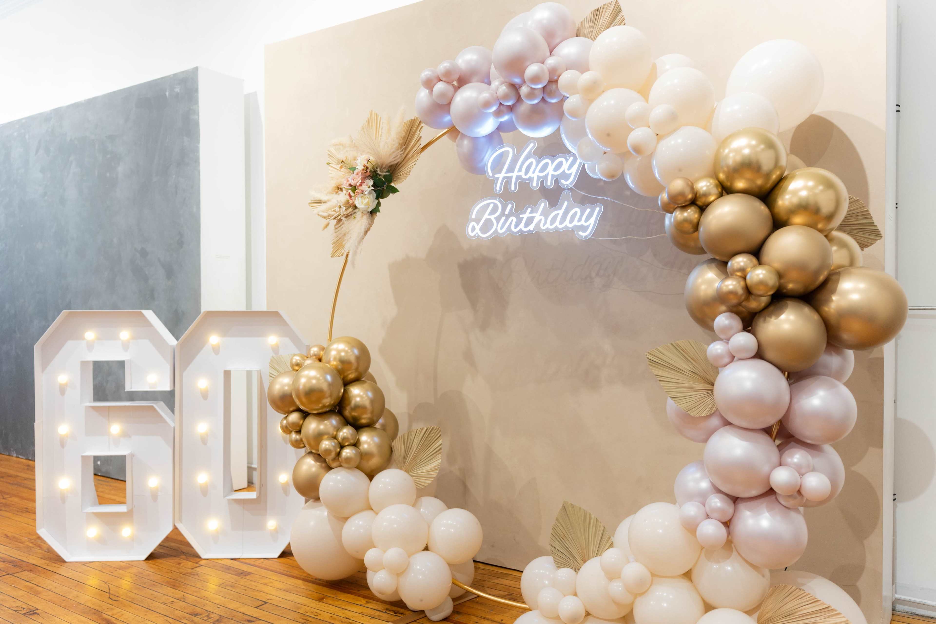The image shows a decorative birthday setup featuring large numerals "60," a balloon arch with white, gold, and beige balloons, and a neon sign that reads "Happy Birthday."