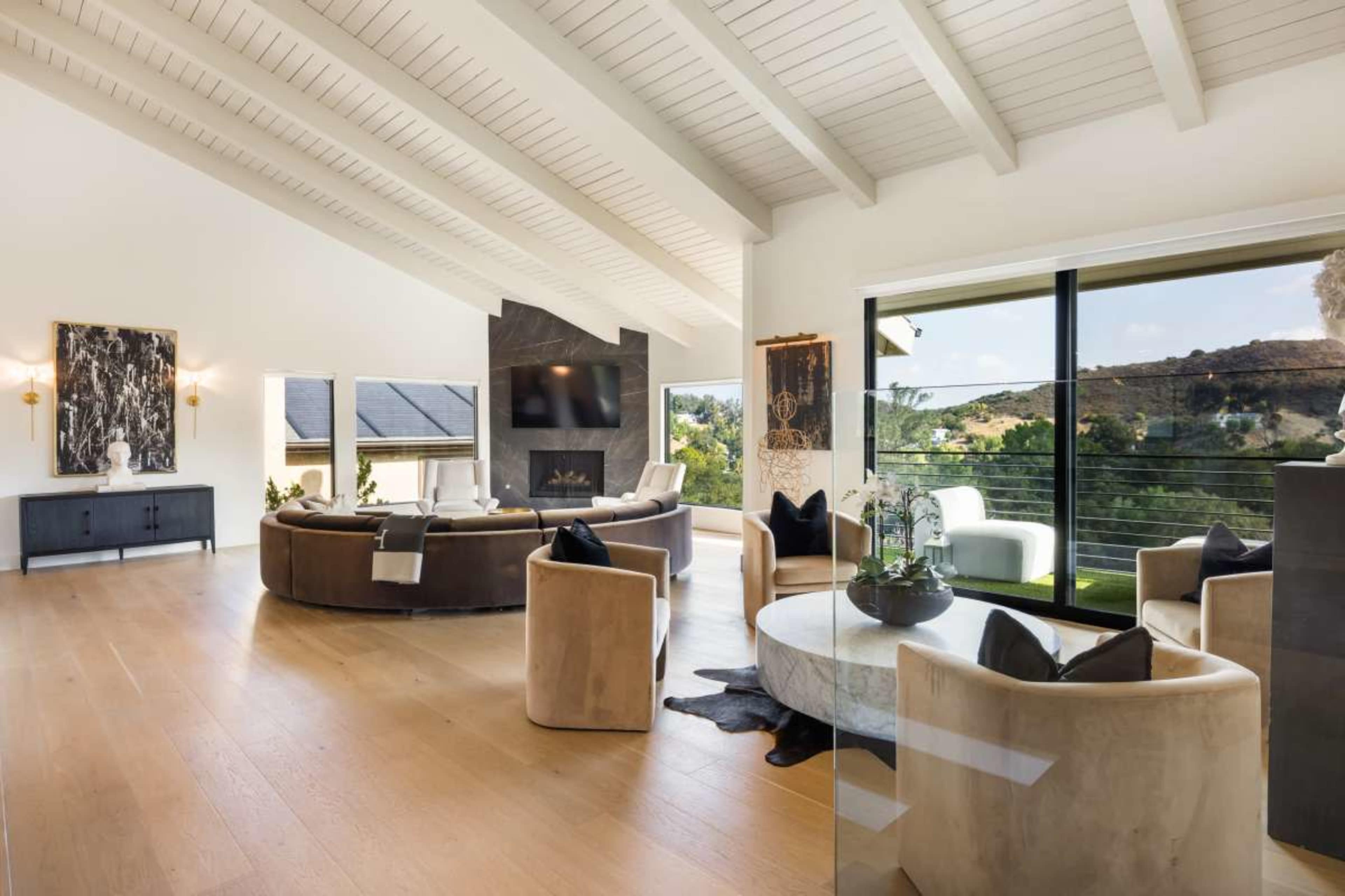The image shows a spacious living room with a high vaulted ceiling, featuring a circular seating arrangement, a large flat-screen television, and large windows overlooking a hillside view.