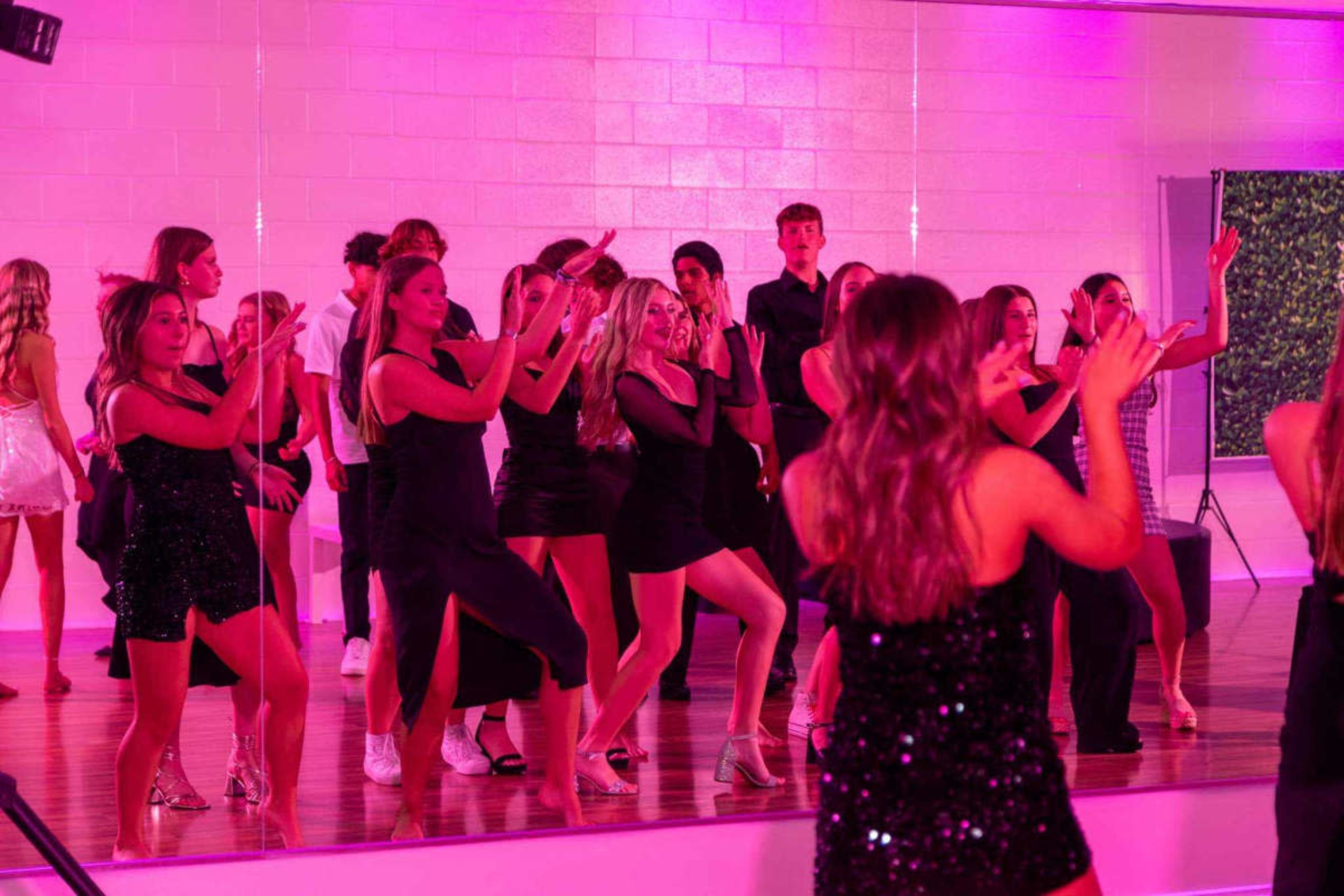 A group of young people dance in front of a large mirror illuminated by pink lights.