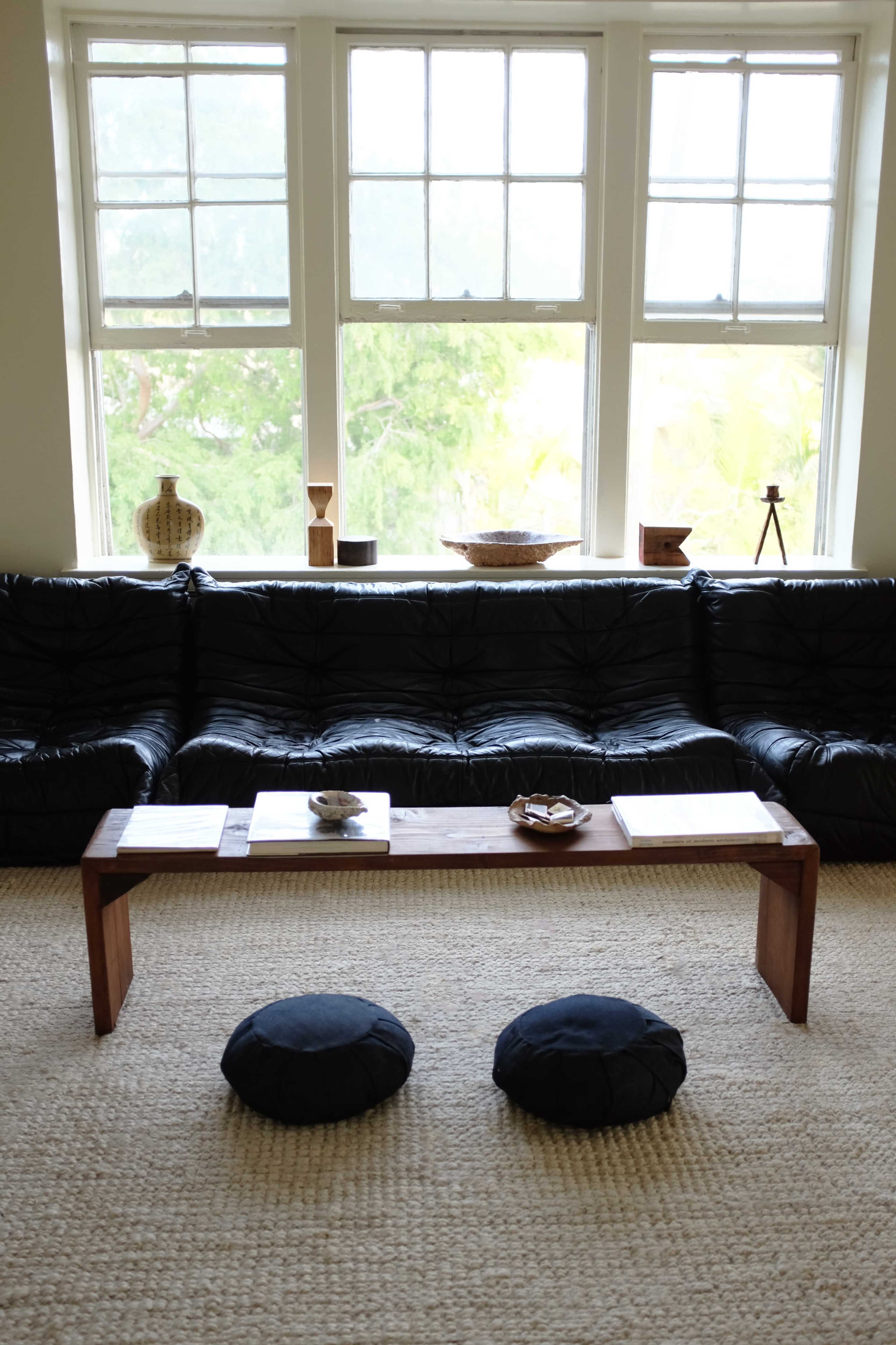 A modern living room features a low wooden coffee table and two round cushions in front of a black couch, with large windows providing natural light.