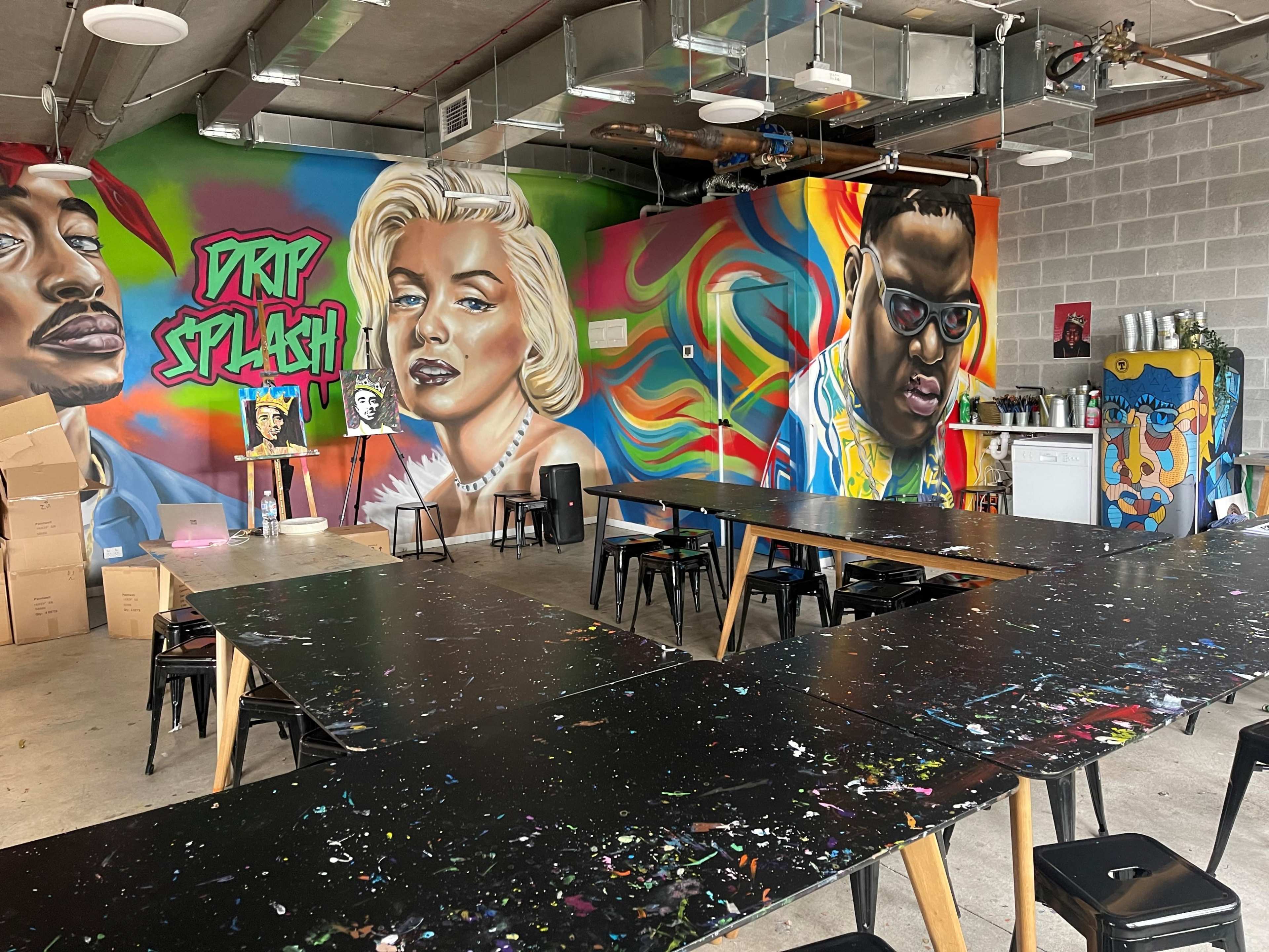 An art studio with several long tables and colorful murals of famous figures on the walls.