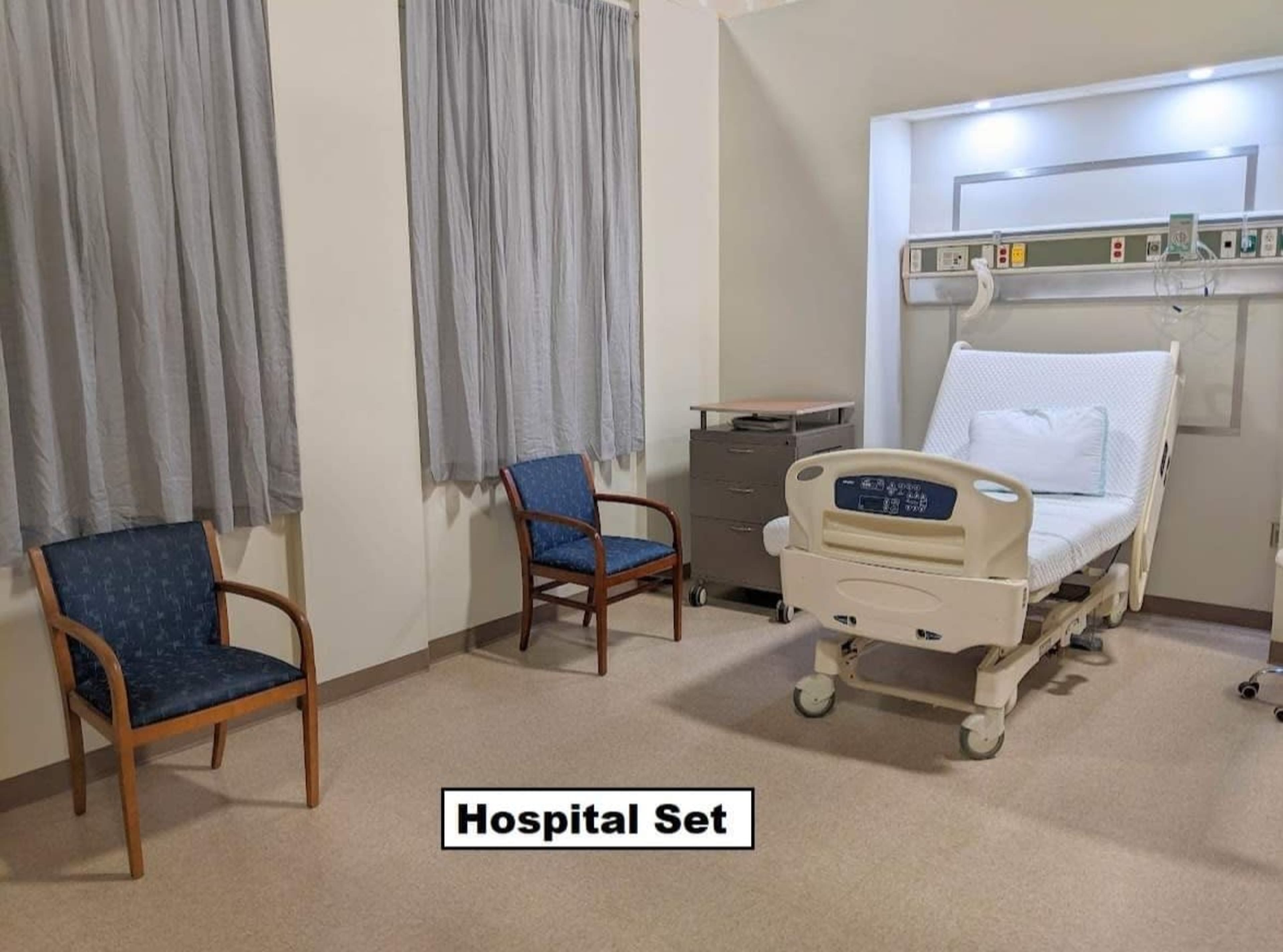The image shows a hospital room featuring a bed with medical equipment, two wooden chairs, and gray curtains.