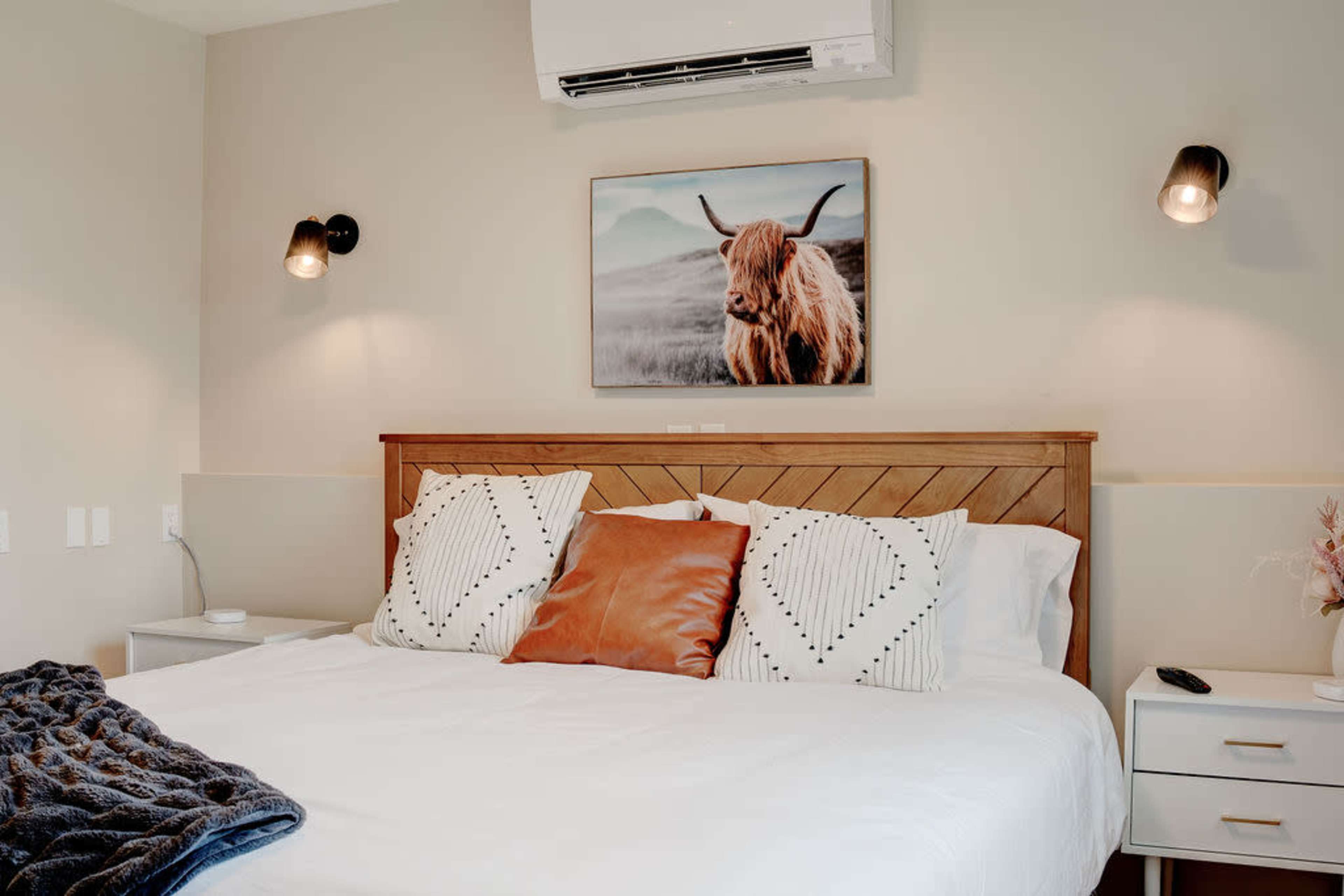 A modern bedroom features a wooden bed with decorative pillows, a wall-mounted air conditioner, and a framed animal portrait above the headboard.