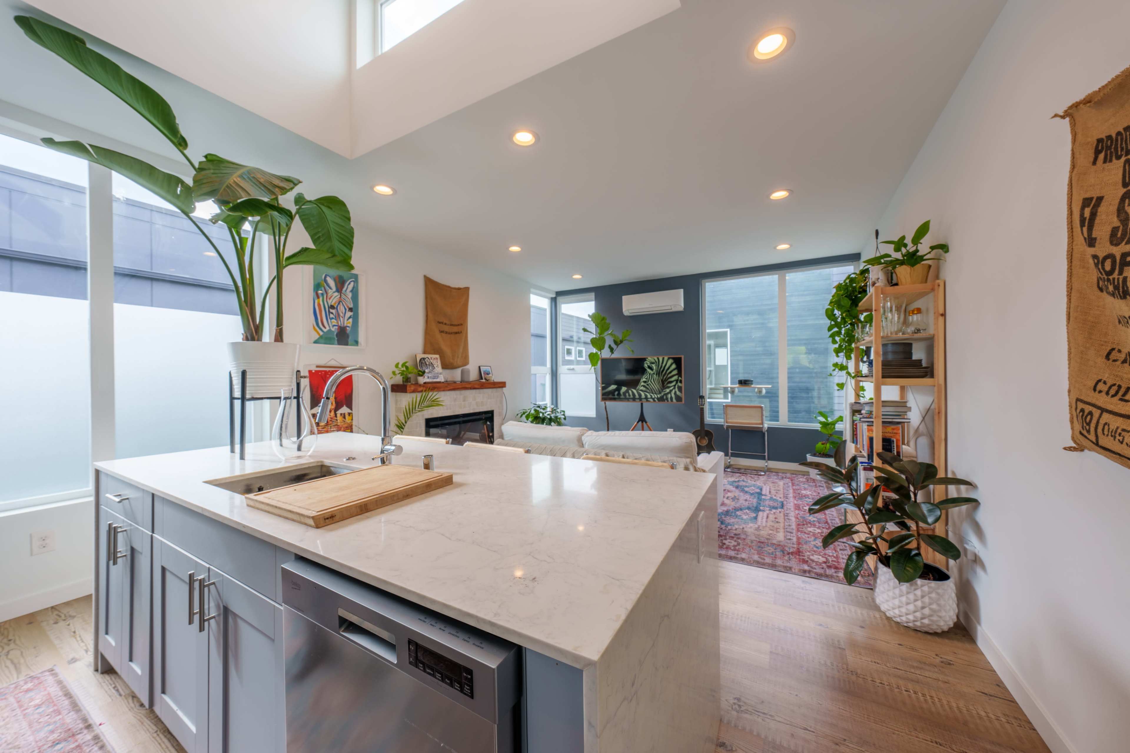 A modern kitchen with a marble countertop, a sink, and various plants placed throughout a bright, open living space.